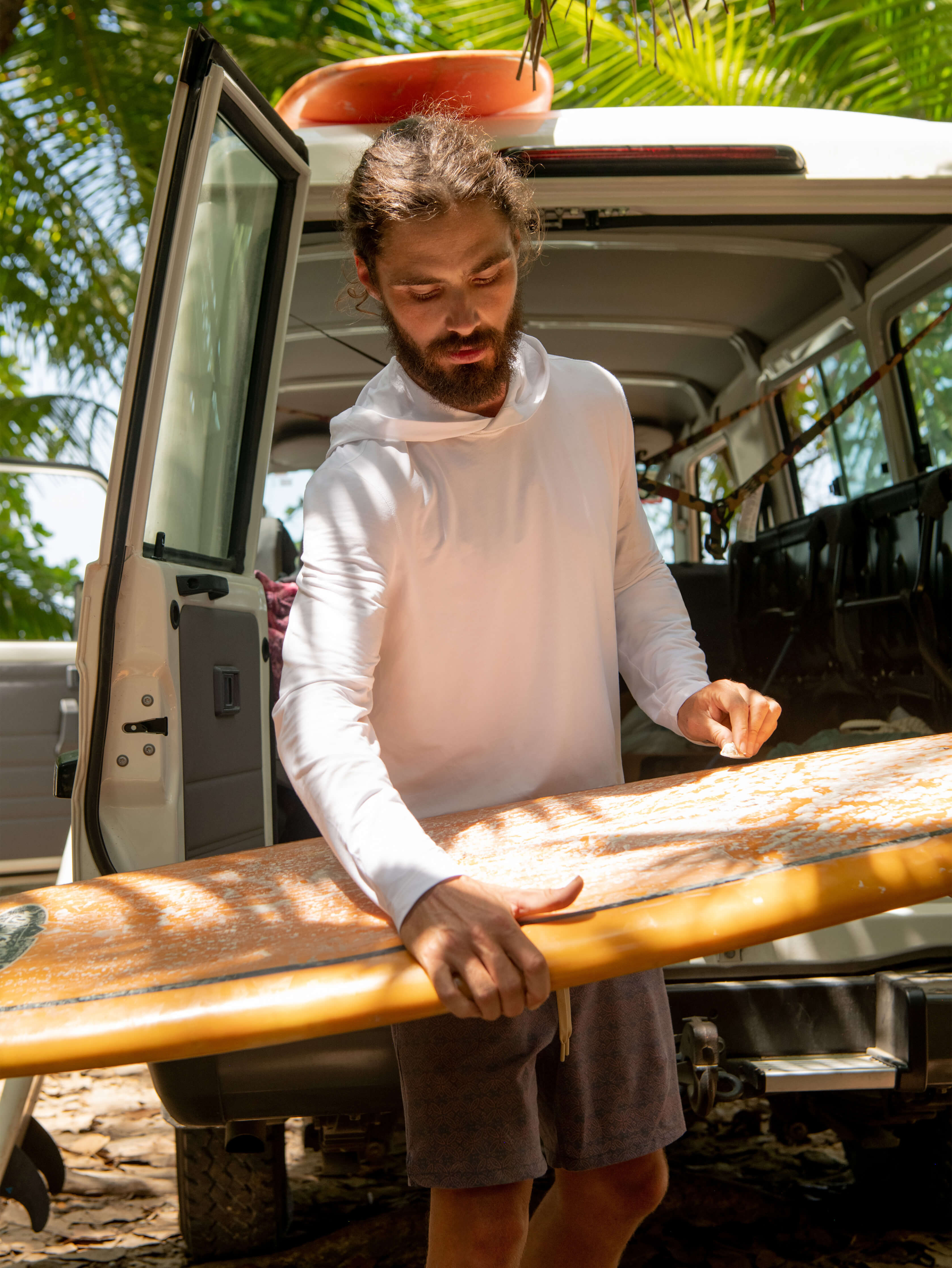 A man standing next to a van, wearing a white hoodie with long sleeves, holding a surfboard.