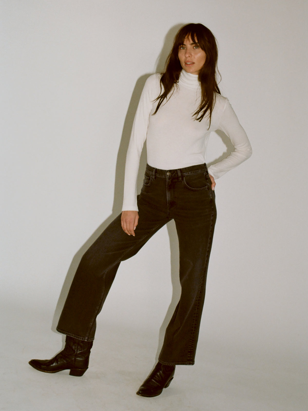 A model wearing a white turtleneck knit with black denim jeans and black boots standing against a plain light gray studio background., image 1 of 7