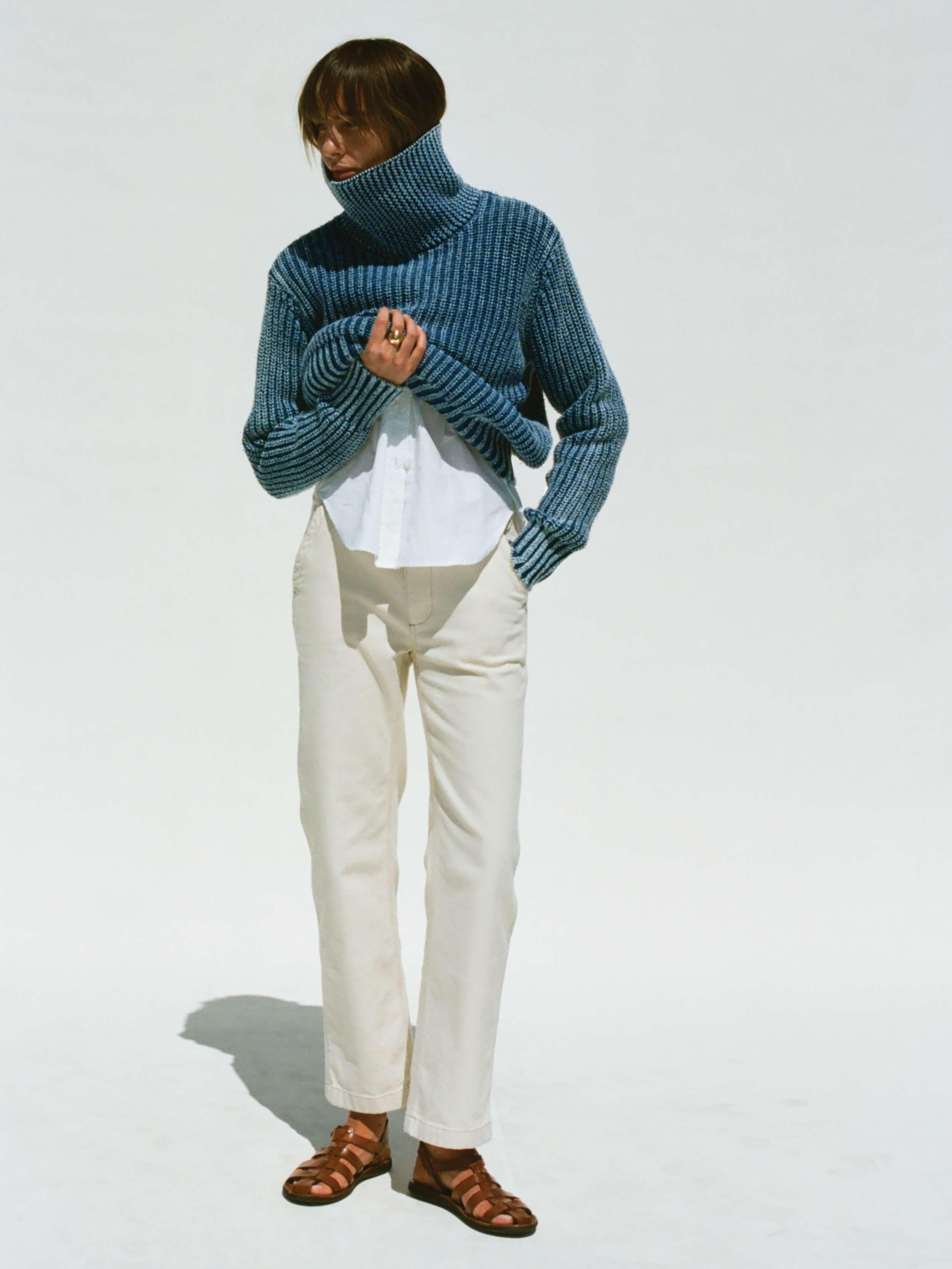 Woman wearing a blue knitted sweater, white pants, and brown sandals on a light gray background