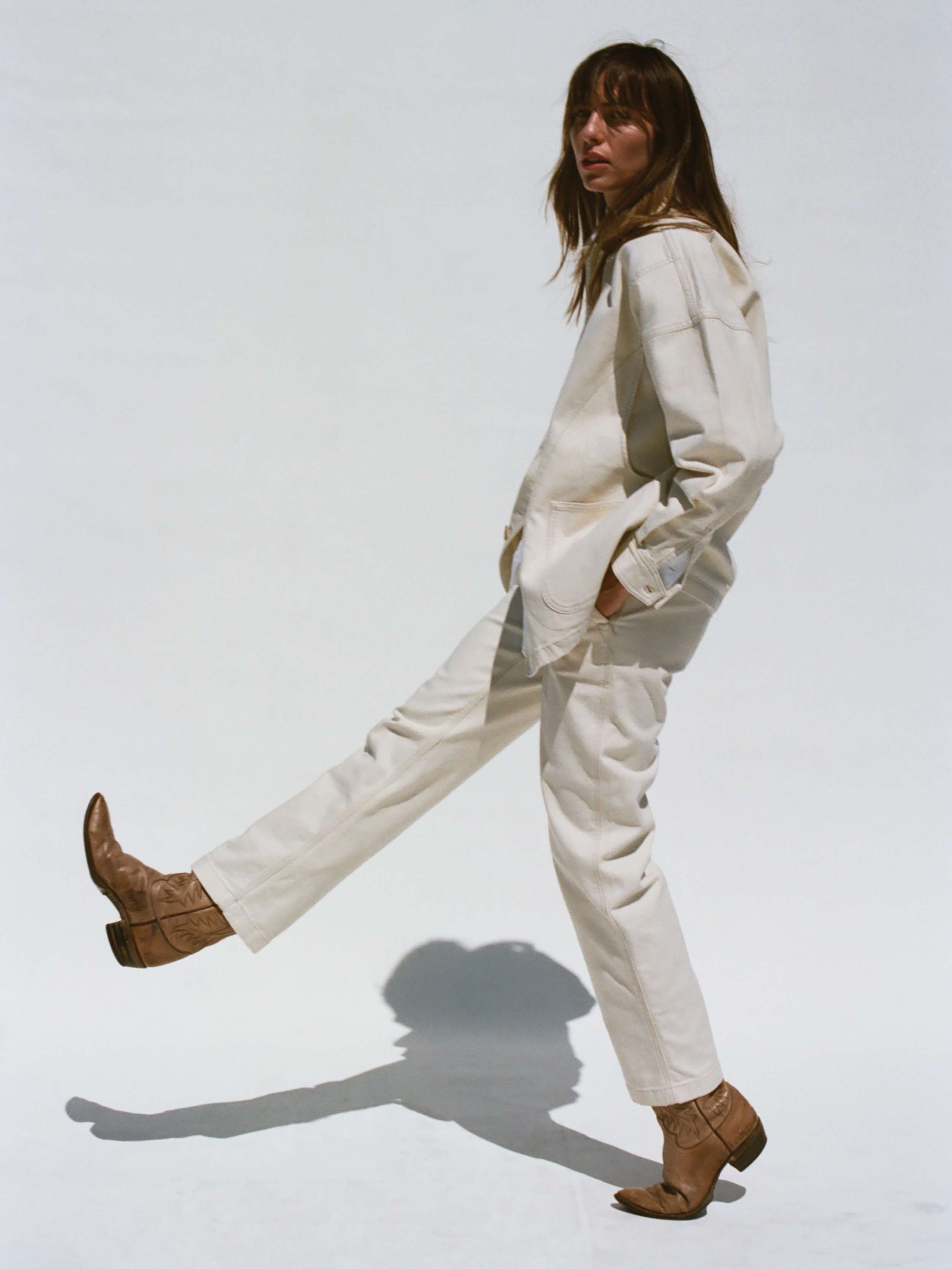 Woman wearing a white jacket and pant set with brown boots on a white background