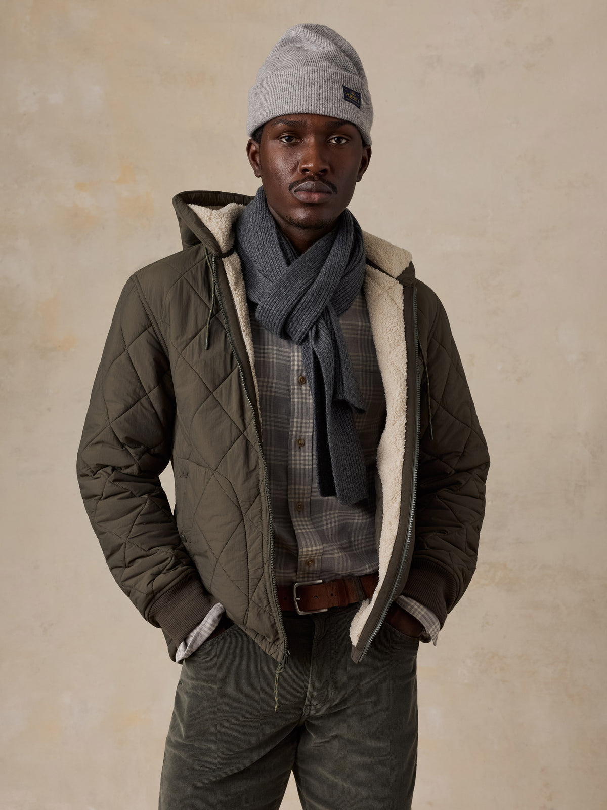 A man wearing a green quilted jacket over a plaid shirt and corduroy pants, image 1 of 5