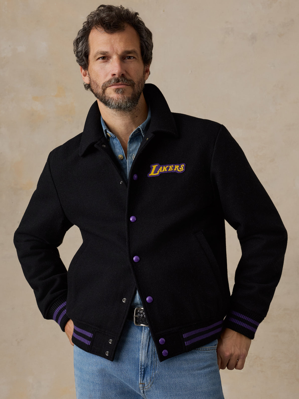 A man wearing a black LA Lakers varsity jacket and blue jeans, image 1 of 8