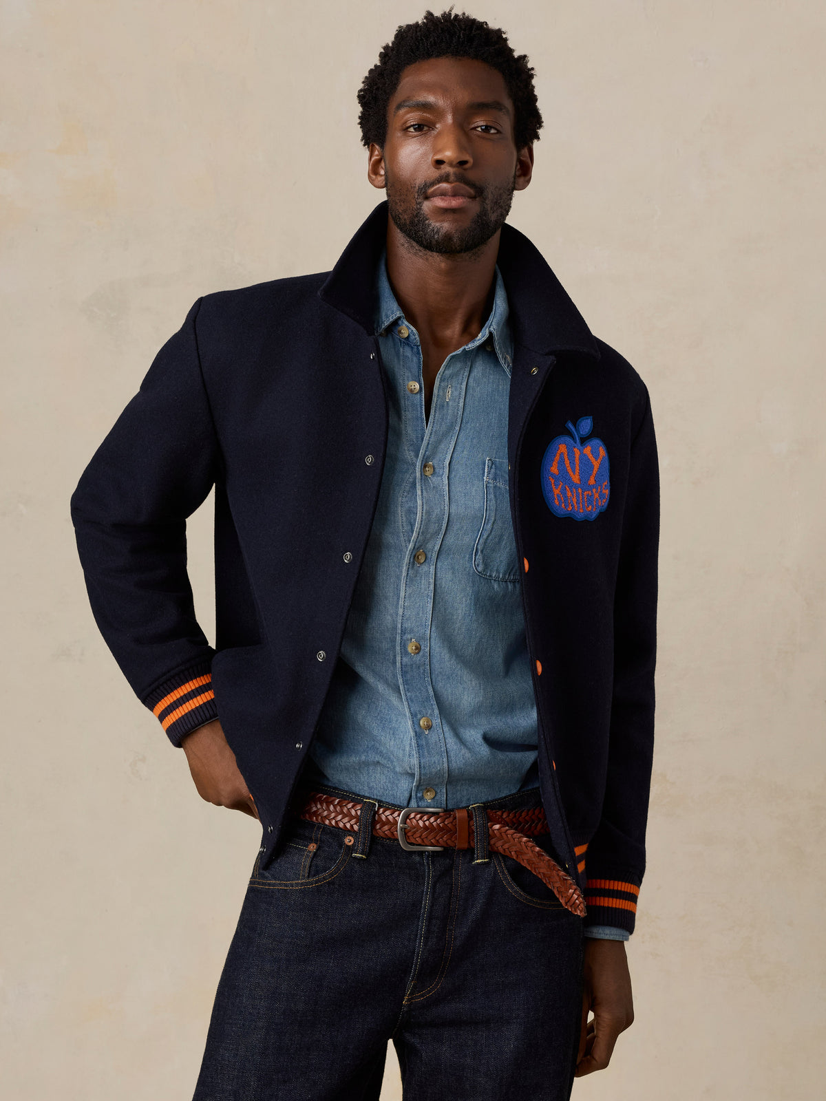 A man wearing a NY Knicks varsity jacket and jeans, image 2 of 8