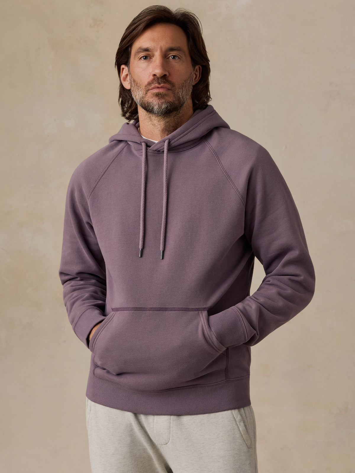 A man wearing a dusty purple pullover hoodie with drawstrings and a front kangaroo pocket, paired with light gray sweatpants, standing against a textured beige wall., image 1 of 5