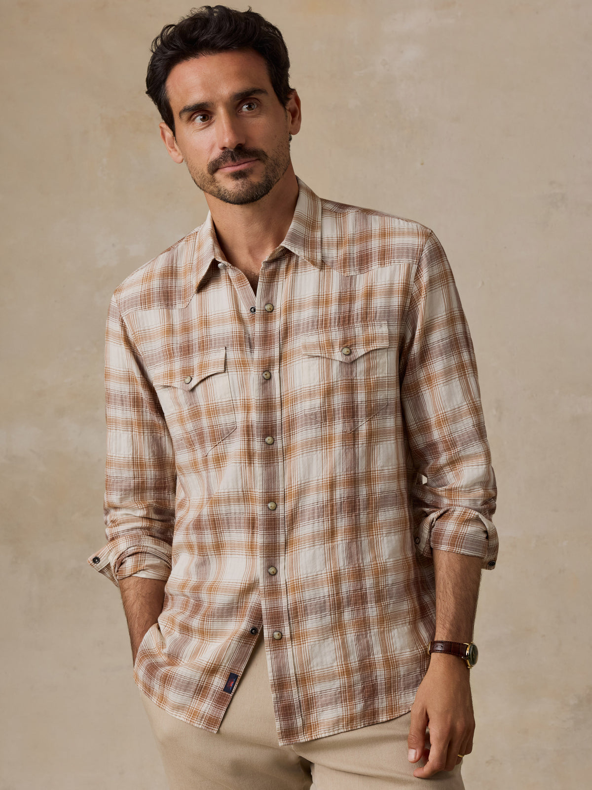 A man wearing a long-sleeve plaid button-up shirt in beige and brown tones, paired with khaki pants, standing against a textured beige background.