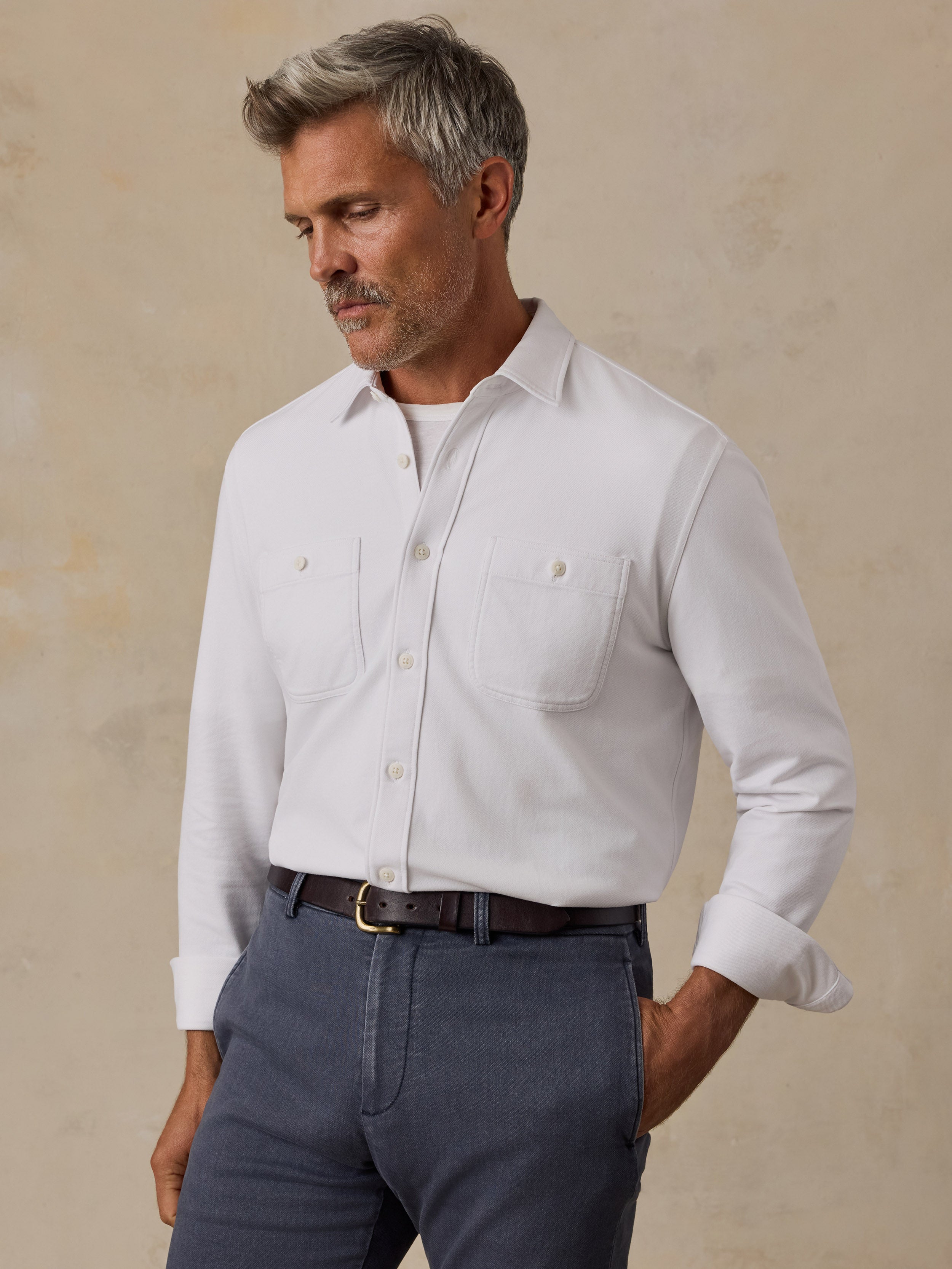 A man wearing a white long-sleeve button-down shirt with two chest pockets, tucked into dark gray trousers with a brown belt, standing against a textured beige background.