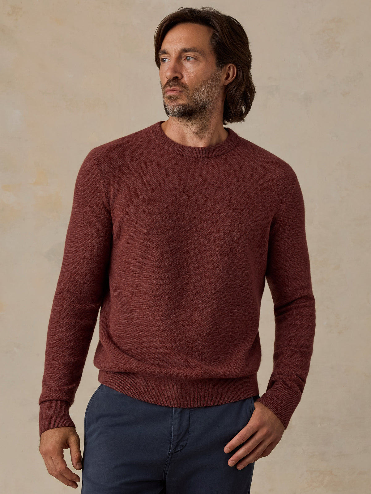 A man wearing a long-sleeve crewneck sweater in a heathered burgundy red, paired with navy blue pants, standing against a textured beige background., image 1 of 5