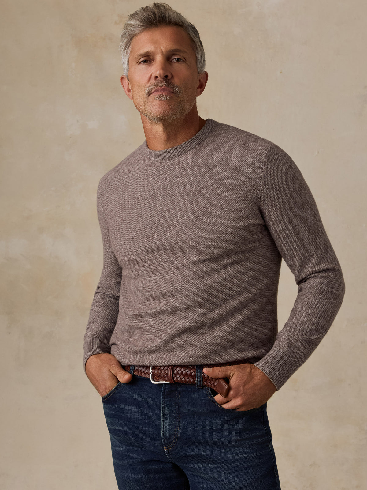 A man wearing a taupe long-sleeve crewneck sweater and dark blue jeans, standing against a textured beige background., image 1 of 5