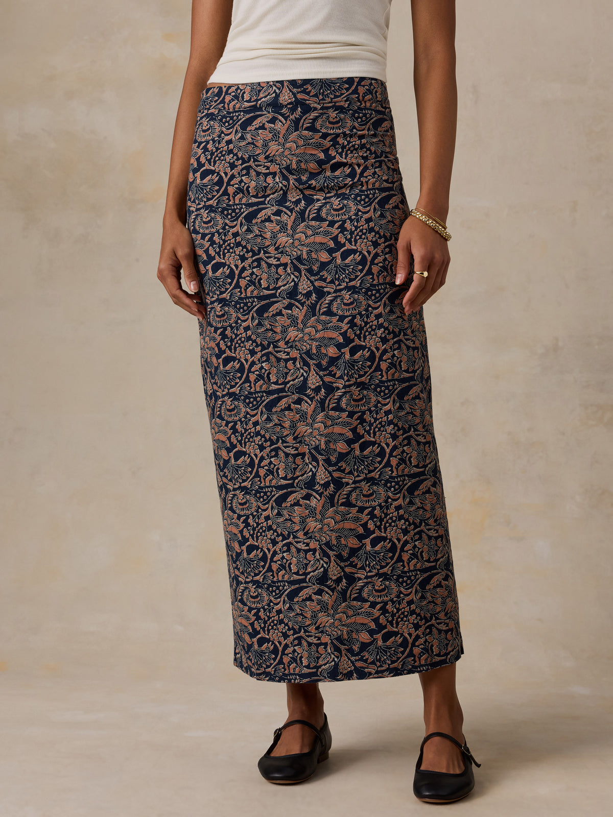 A woman wearing a long navy blue skirt with an intricate tan floral damask pattern, paired with a white top and black flats, standing against a textured beige studio backdrop., image 2 of 6