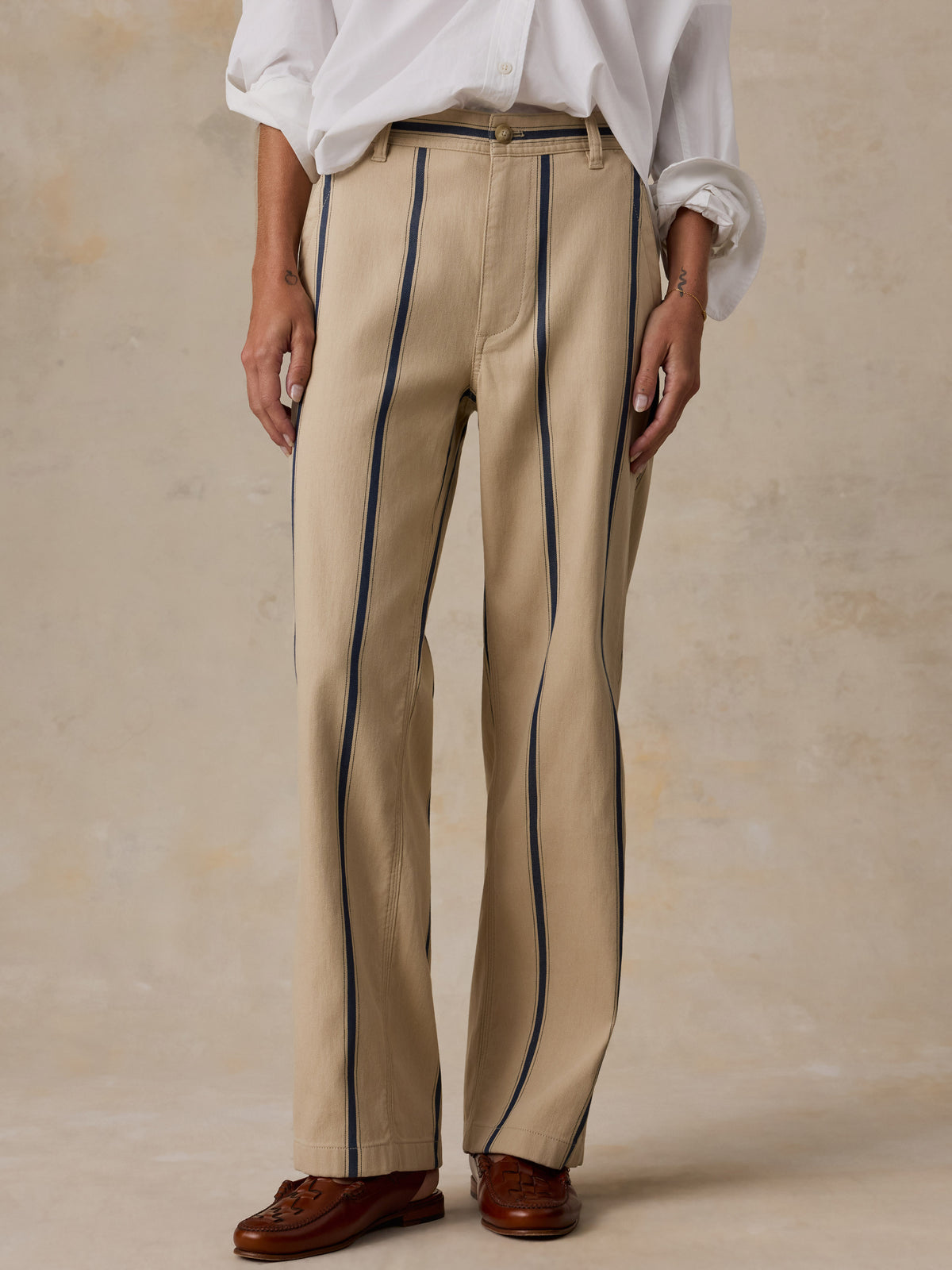 A person wearing beige wide-leg trousers with dark blue vertical pinstripes, styled with a white shirt and brown leather shoes against a textured beige studio backdrop., image 2 of 8