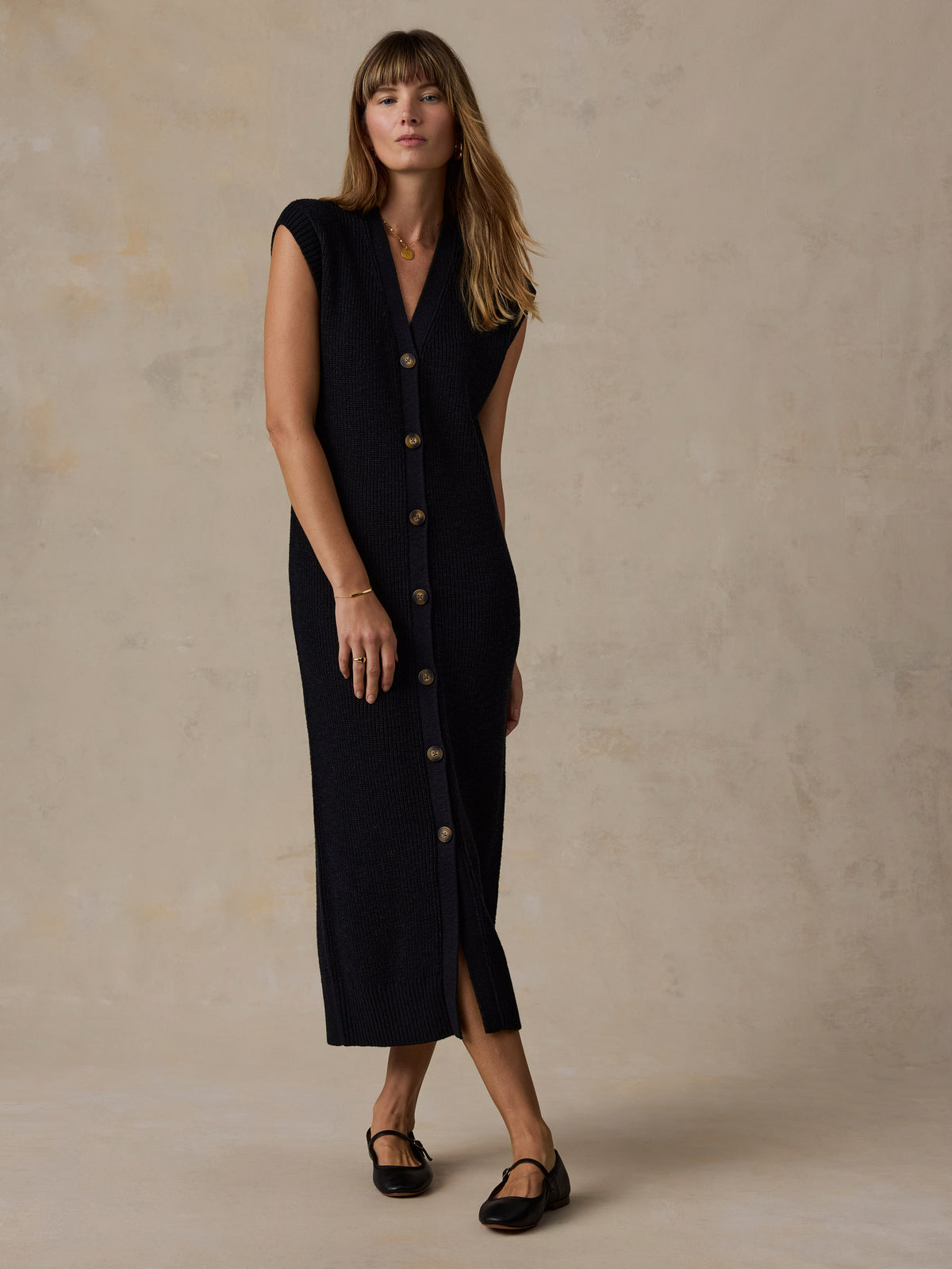 A woman modeling a sleeveless, dark navy midi dress with a front button closure, standing against a textured beige studio backdrop., image 3 of 5