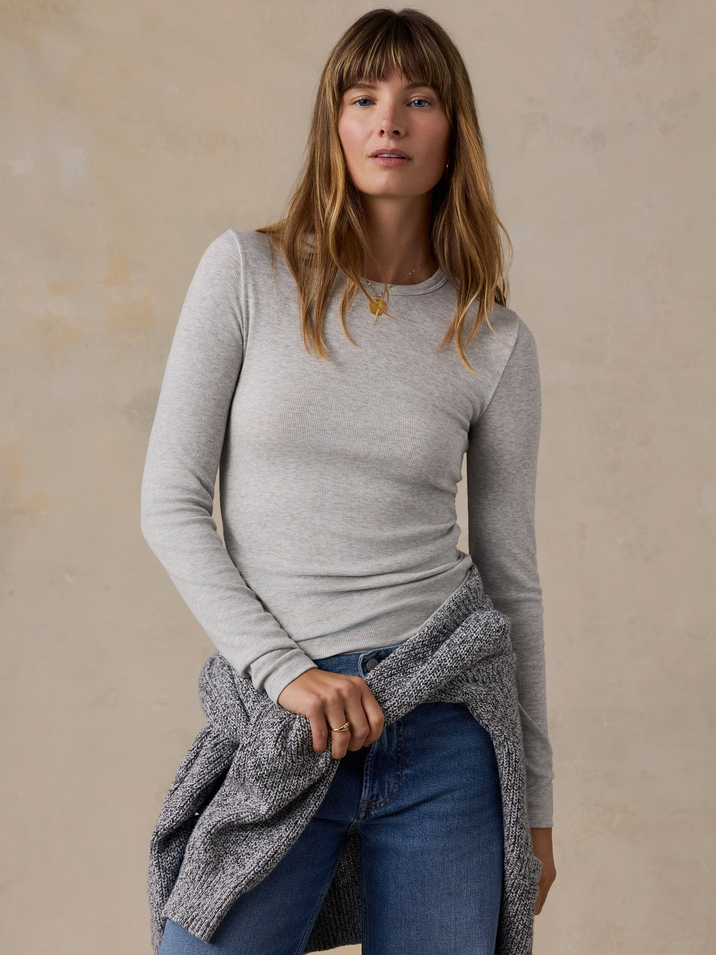 A woman modeling a light heather gray long-sleeve crewneck top, paired with blue jeans and a gray knit sweater draped off her shoulders, against a textured beige studio backdrop.