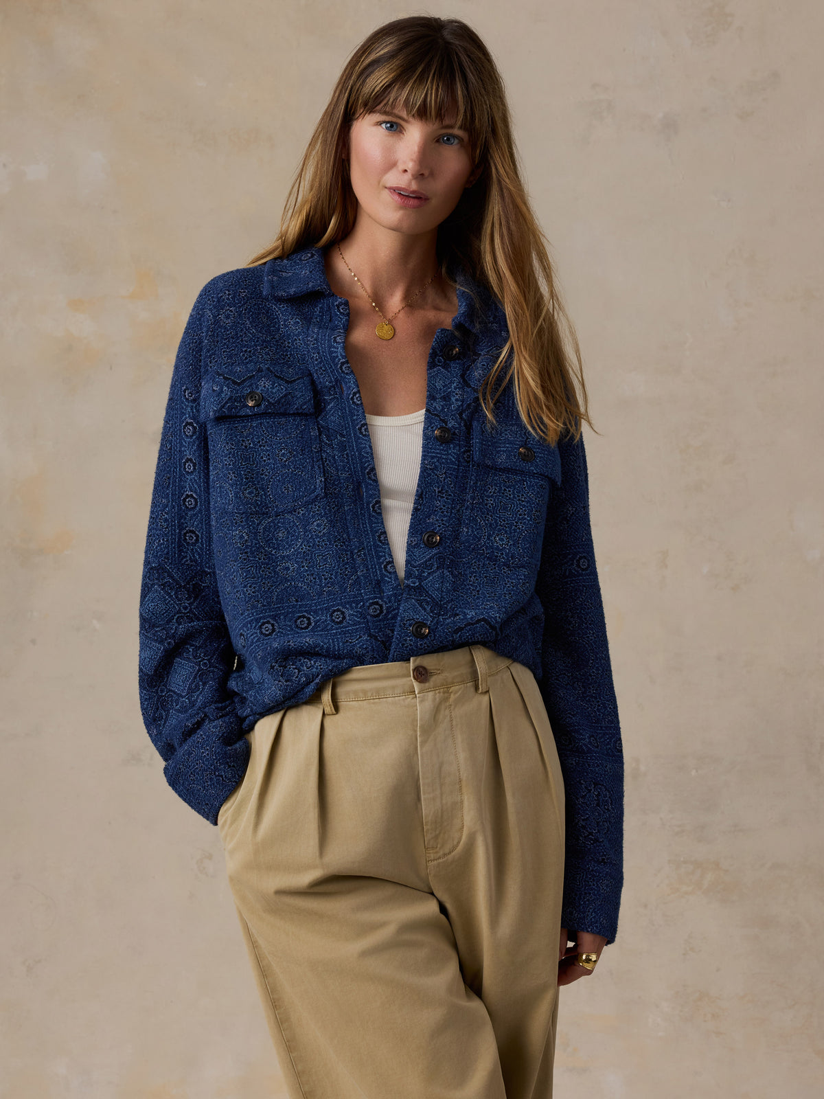 A woman wearing a dark blue patterned button-up shirt jacket over a white top and beige pleated trousers, standing against a textured beige wall., image 1 of 7