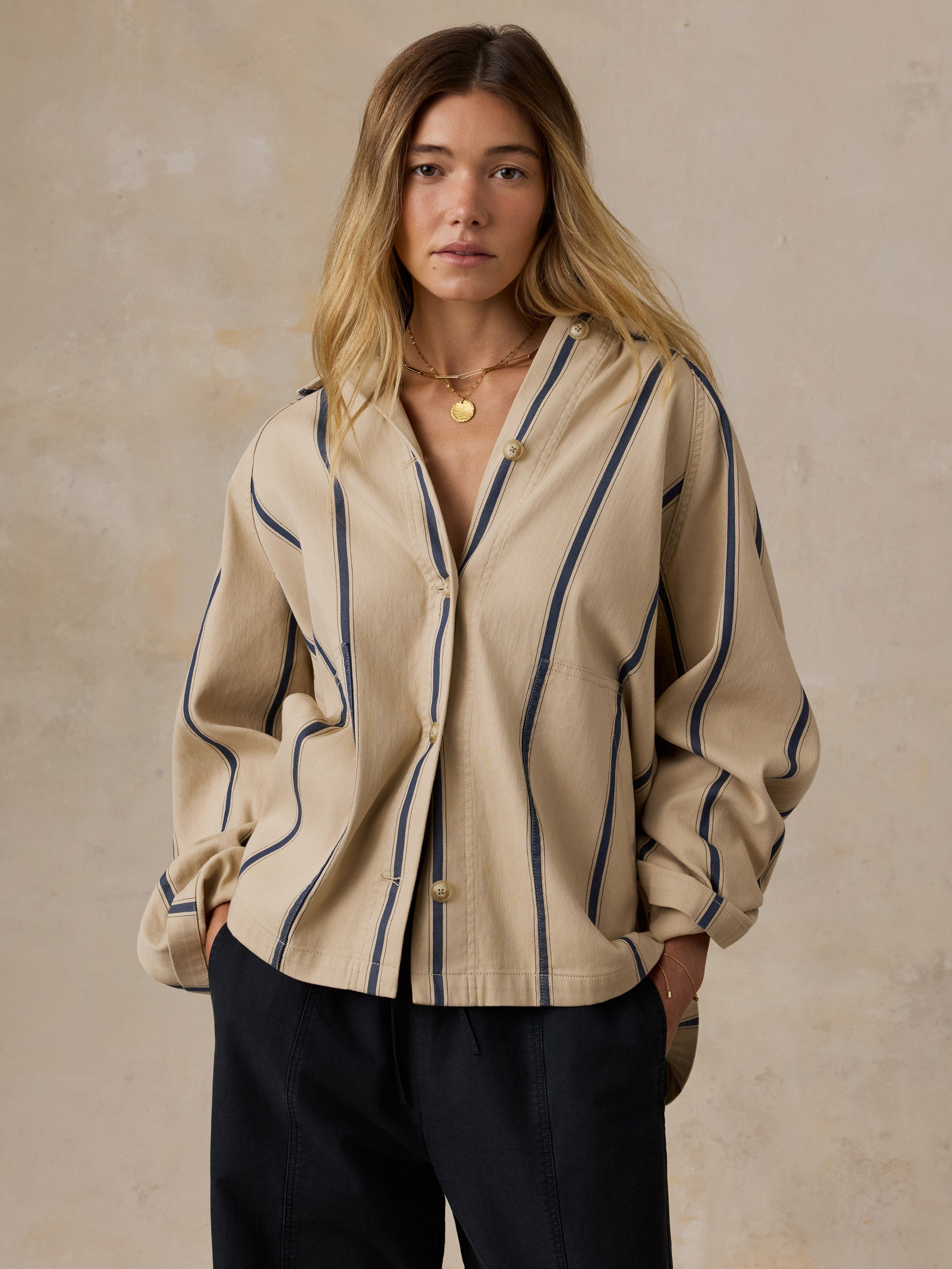 A woman wearing a beige button-front shirt with vertical navy stripes and dark pants, standing against a textured beige wall.