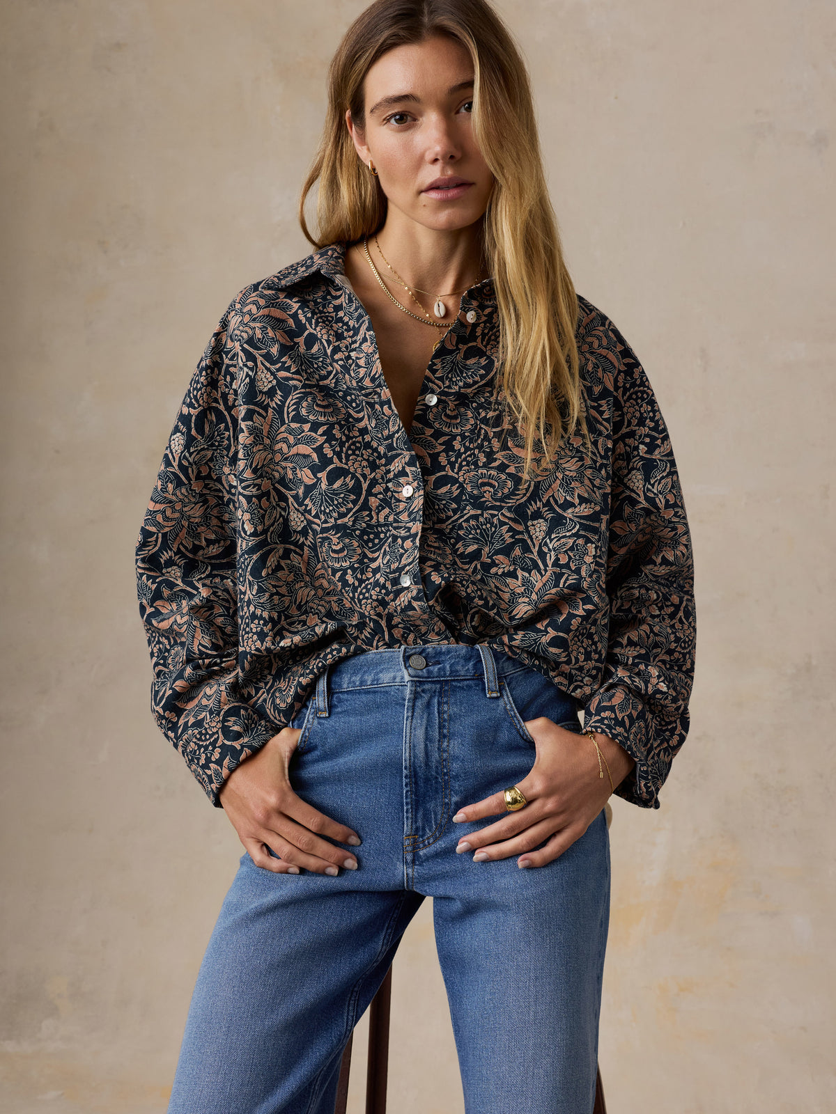 A woman wearing a dark navy and brown floral print button-up shirt tucked into blue jeans, standing against a textured beige wall., image 2 of 7