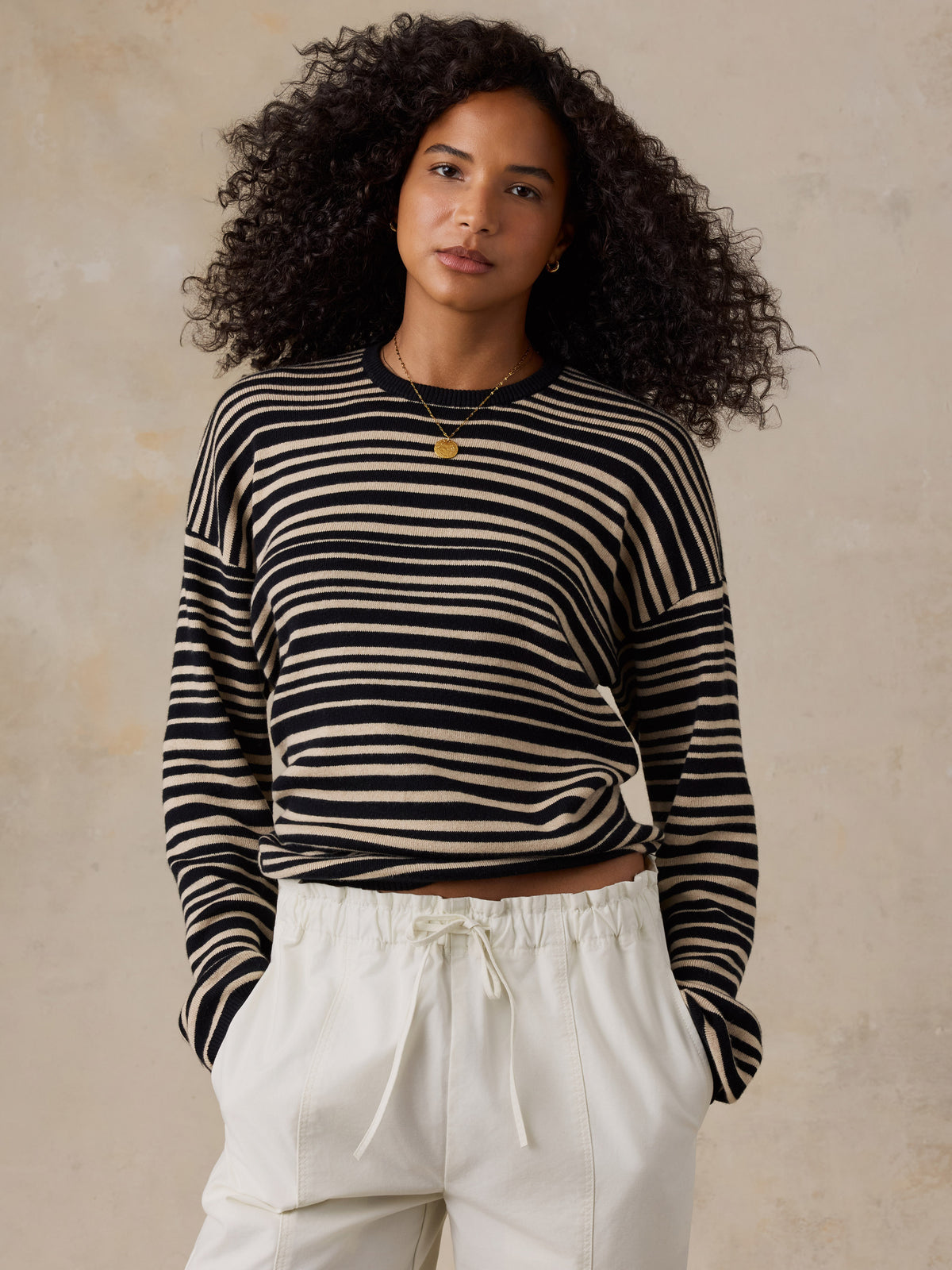 A woman with curly dark hair wearing a black and beige horizontally striped long-sleeve knit top and cream drawstring pants, standing against a textured beige wall., image 3 of 6