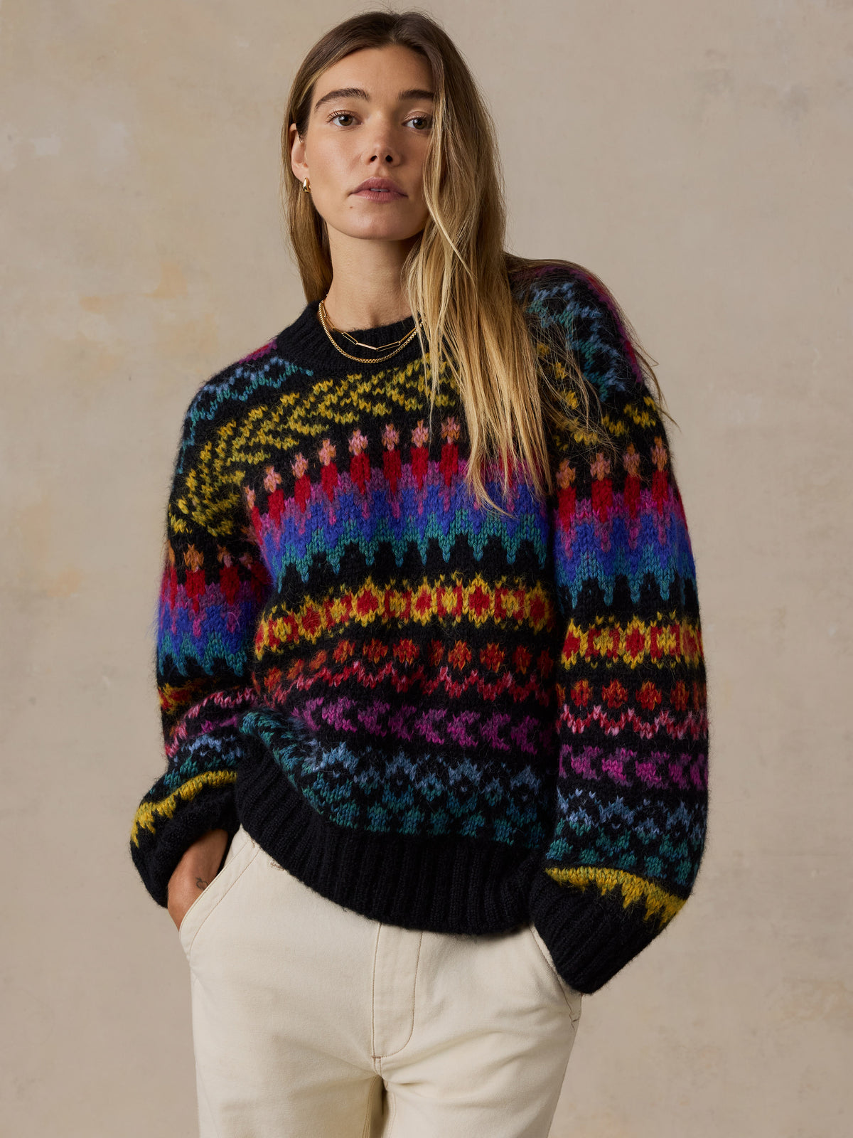 A woman wearing a colorful Fair Isle knit sweater with horizontal stripes of red, orange, yellow, green, blue, and purple on a black base, paired with cream pants, set against a textured beige studio background., image 6 of 7