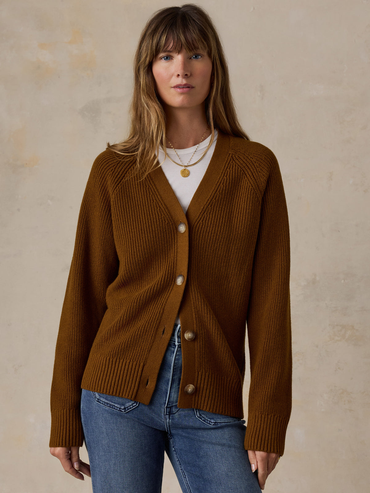 A woman wearing a ribbed brown cardigan over a white top and blue jeans, standing against a textured beige wall., image 1 of 5