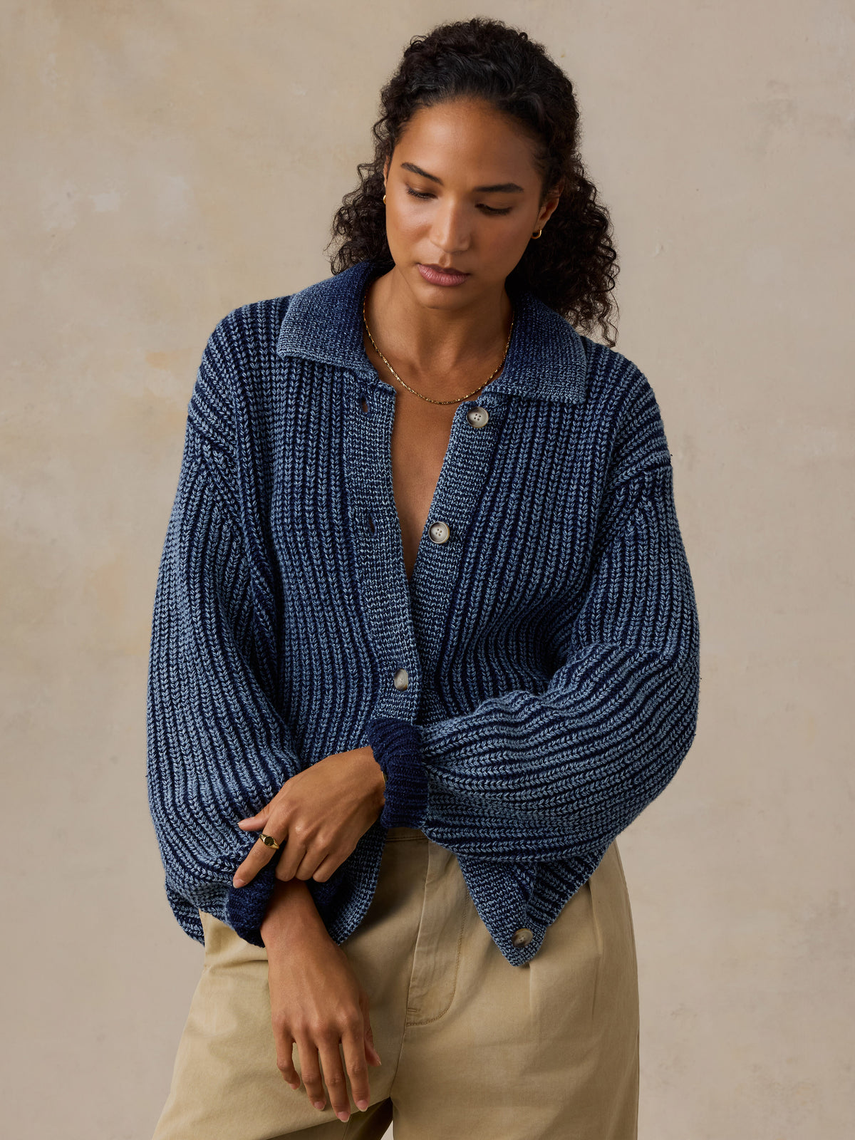 A woman wearing a textured navy blue knit button-up cardigan with voluminous sleeves and beige pants, standing against a neutral textured beige wall., image 6 of 8