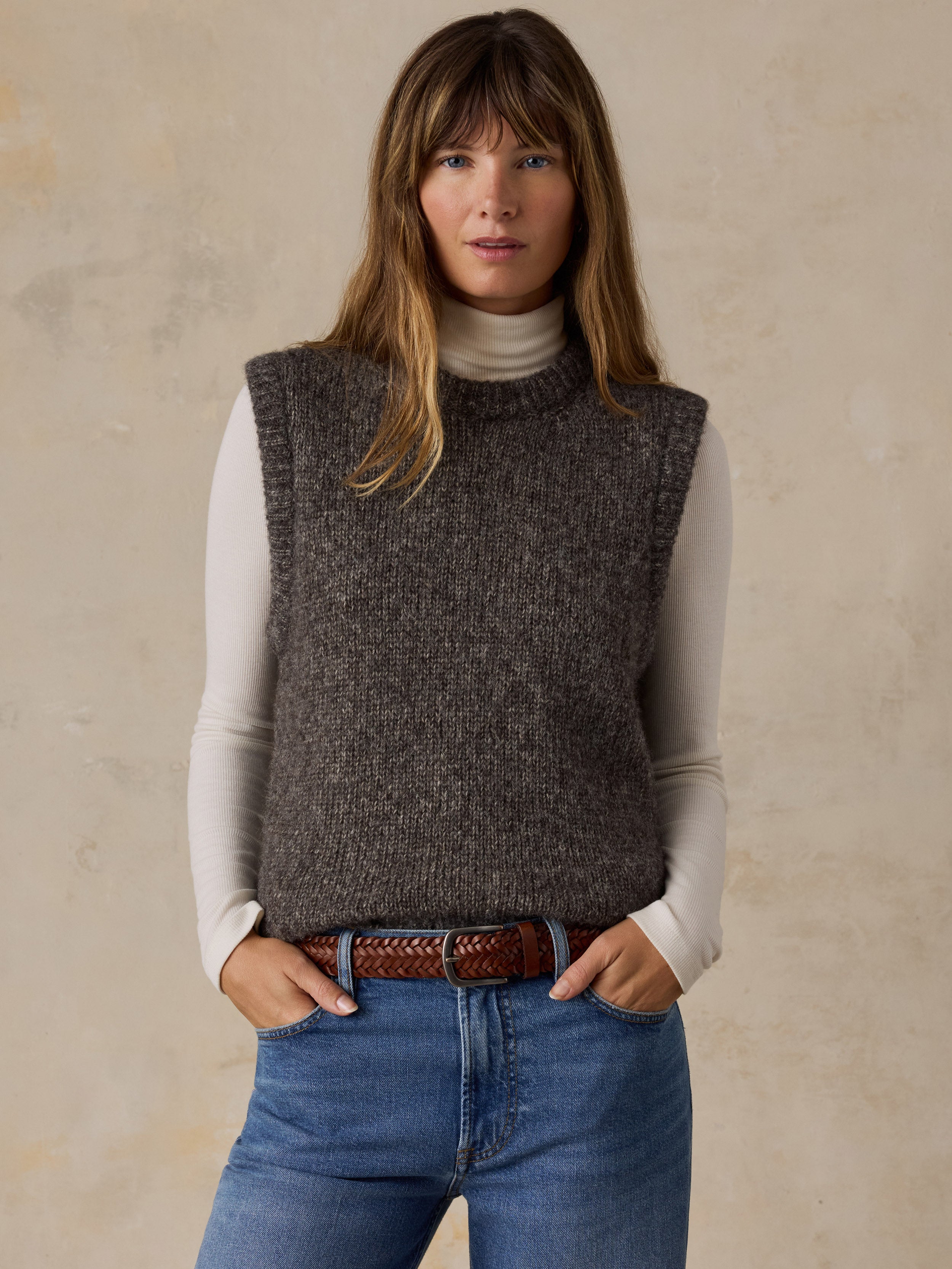 A woman wearing a grey sweater vest with a white turtleneck and blue jeans