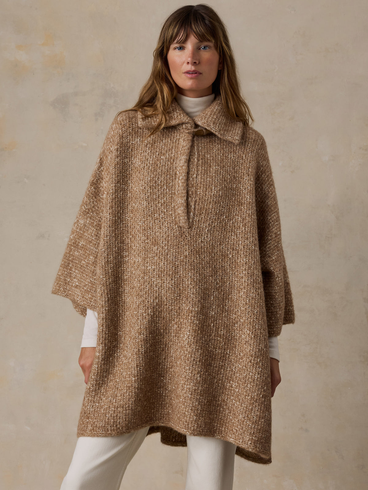 A woman wearing a beige sweater poncho over a white turtleneck and white pants, image 2 of 7
