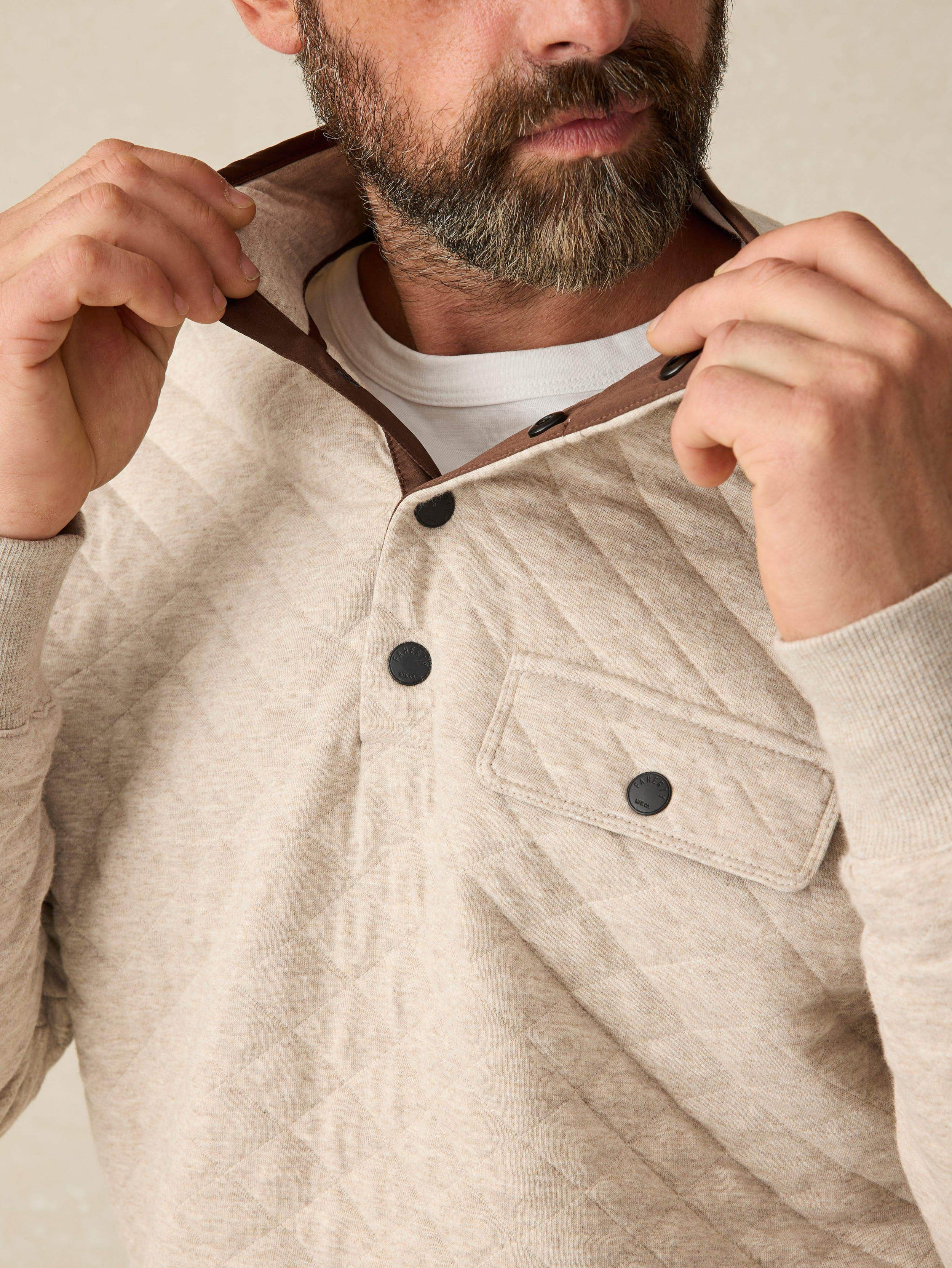 Man wearing an oatmeal quilted pullover sweater with button left front chest pocket against a beige background