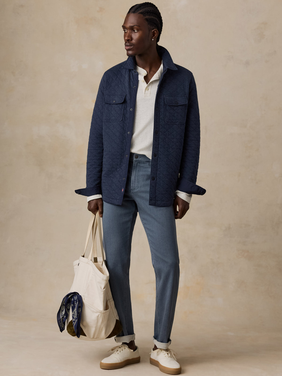 Man wearing a navy quilted jacket with blue jeans on a beige background, image 6 of 7