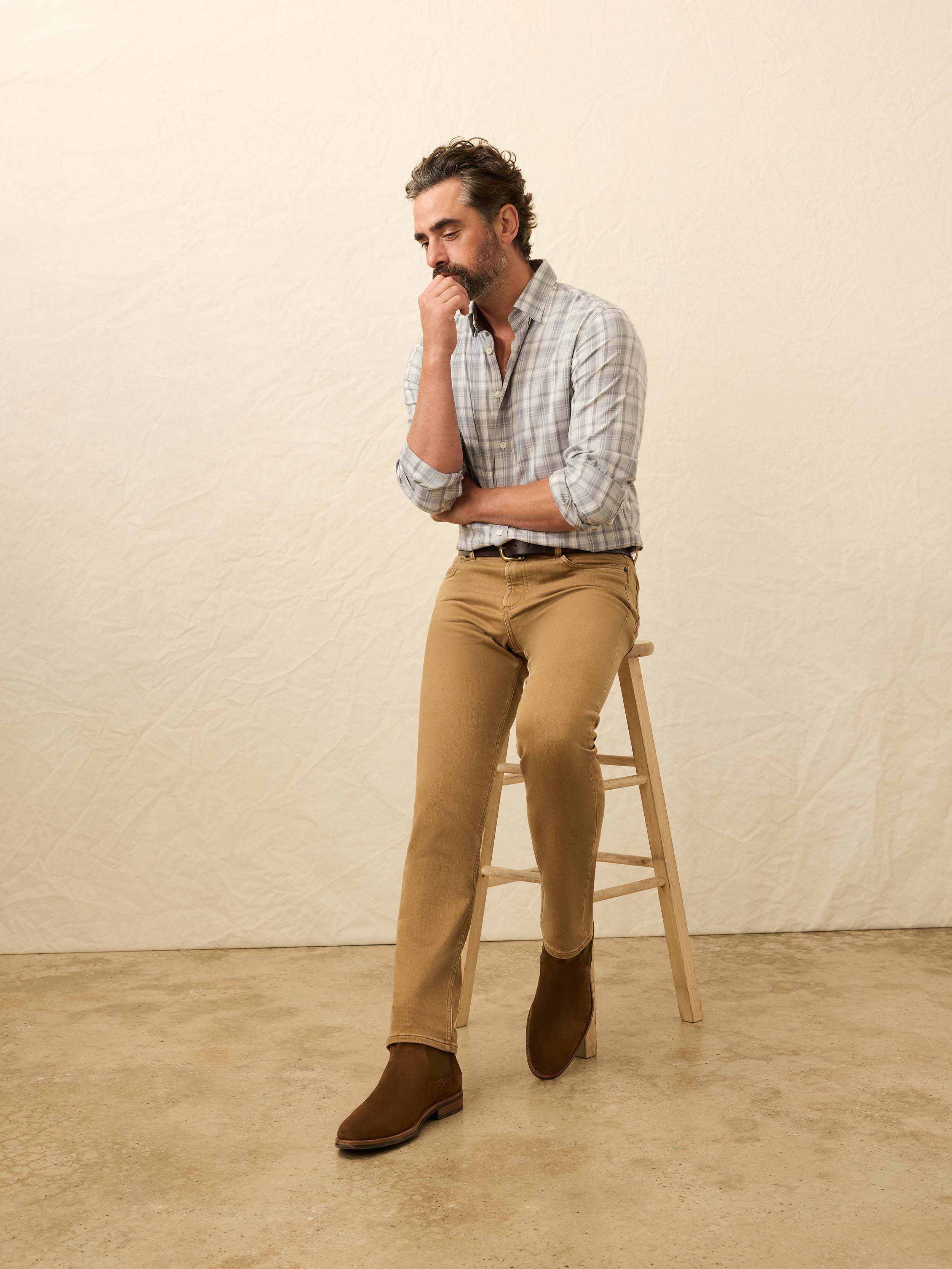 Man sitting on a stool wearing a plaid shirt, light brown pants, and brown shoes against a beige background