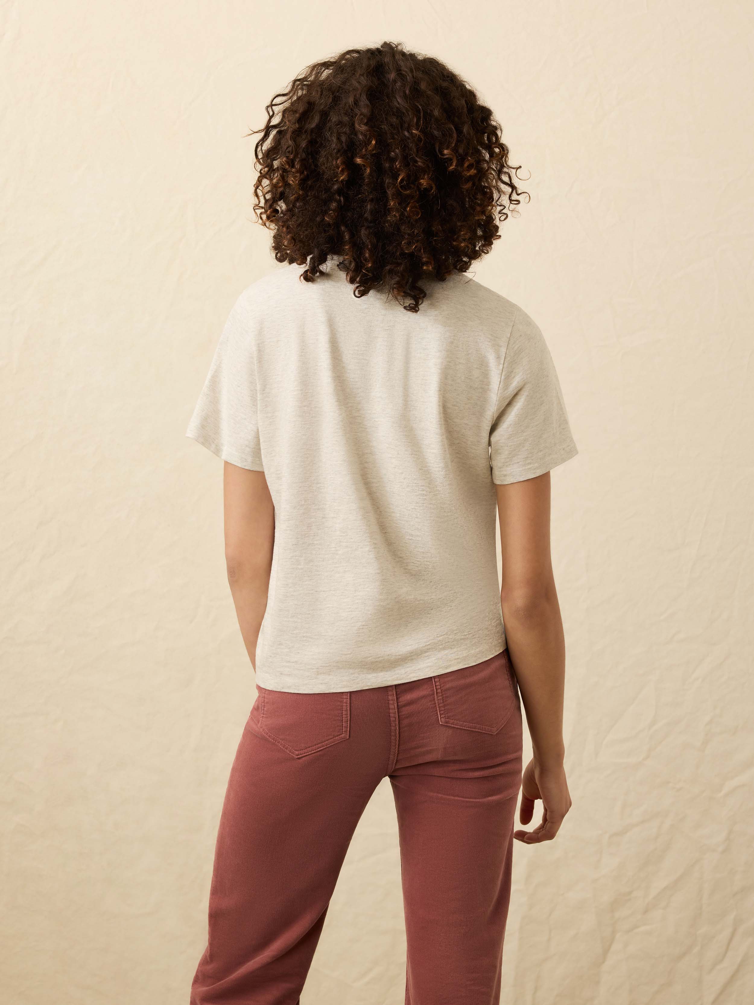 Back view of a woman wearing a light heather grey t-shirt and pink corduroy pants against a beige background