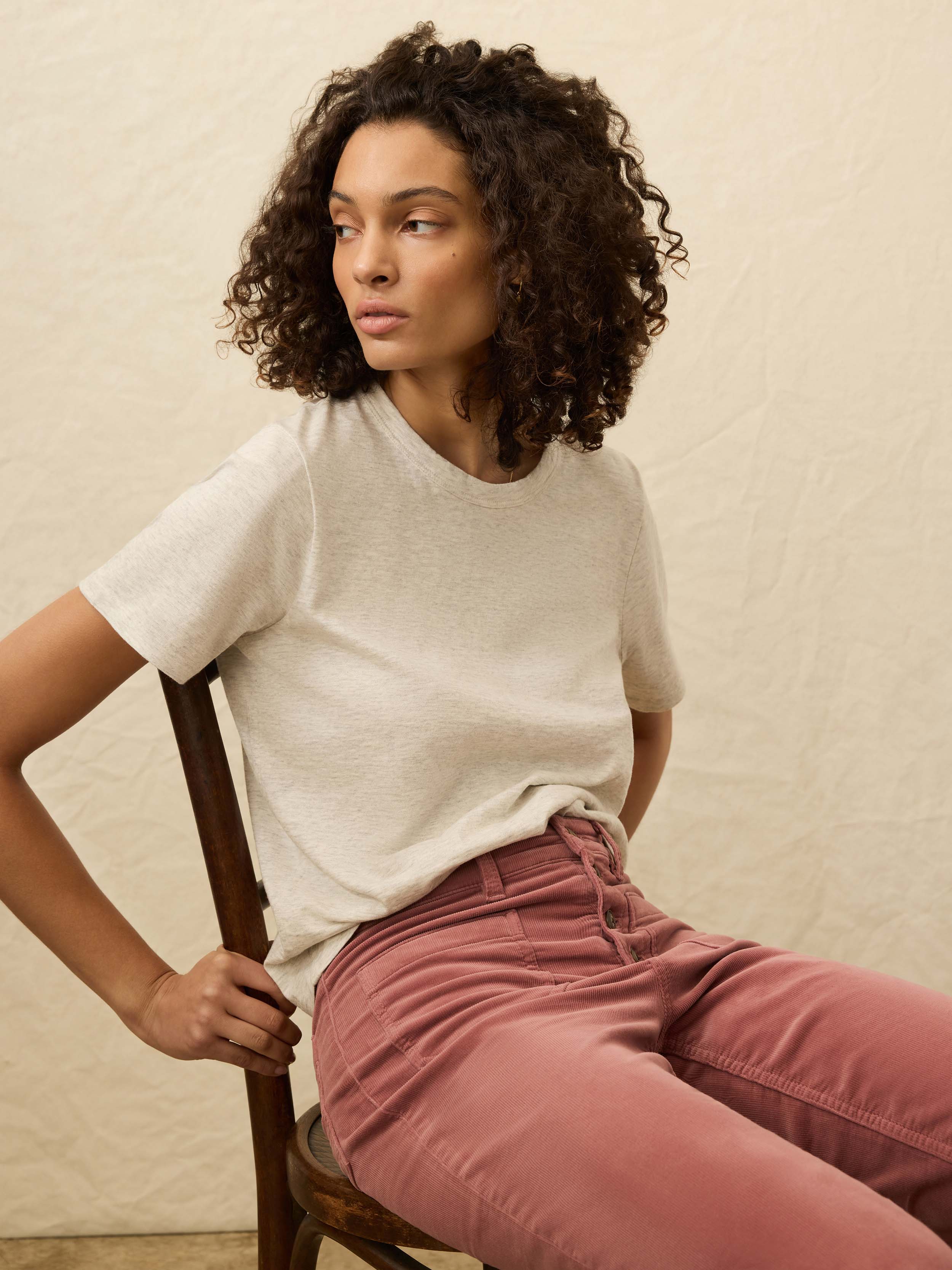 Woman sitting on a chair wearing a light heather grey t-shirt and pink corduroy pants against a beige background