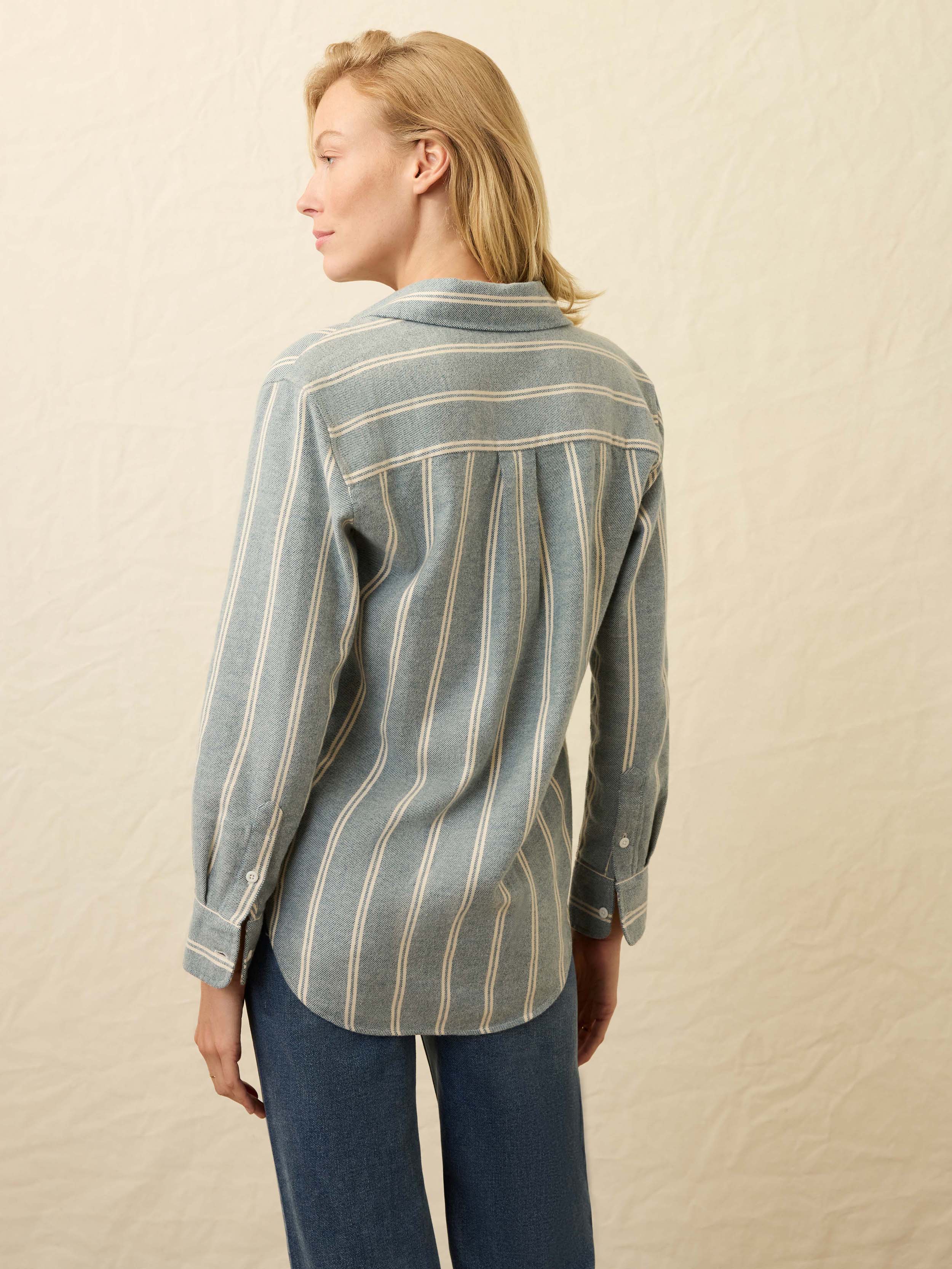 Back view of a woman wearing a light blue and white striped denim shirt with blue jeans against a beige background