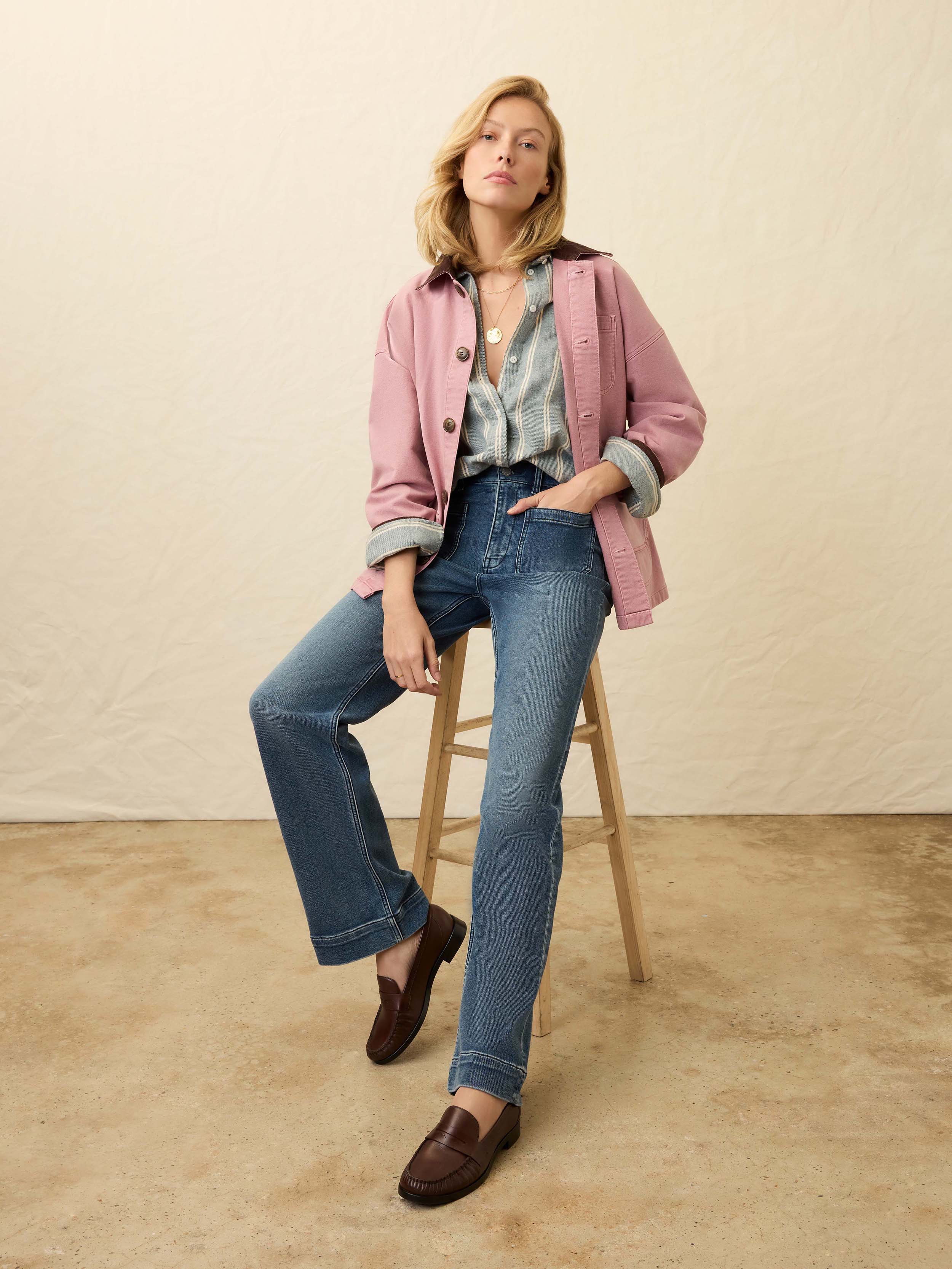 Woman wearing a pink jacket over a light blue and white striped denim shirt with blue jeans against a beige background