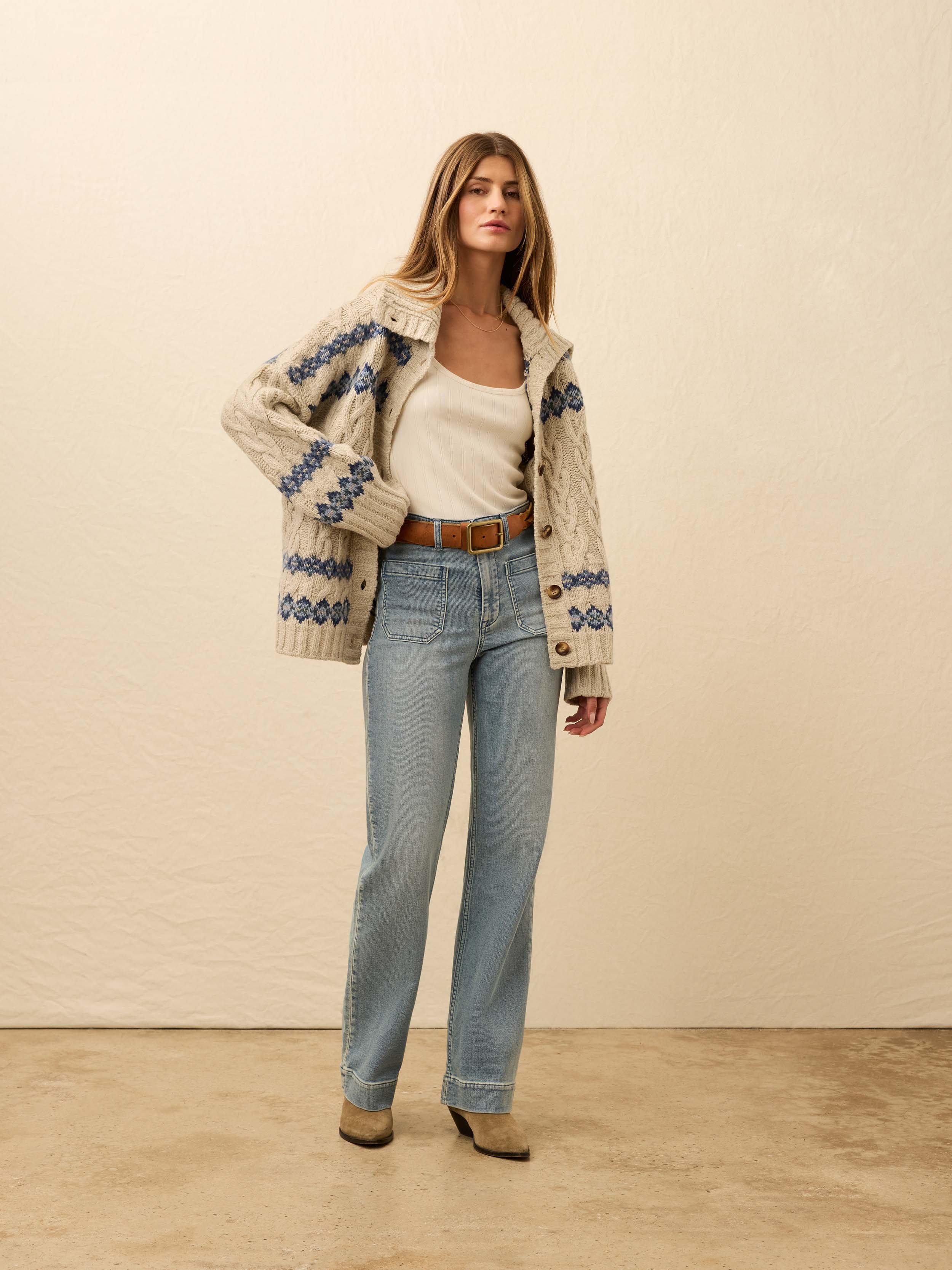 Woman wearing a cream-colored knit cardigan with blue fairisle pattern, light blue jeans, and brown boots against a beige background