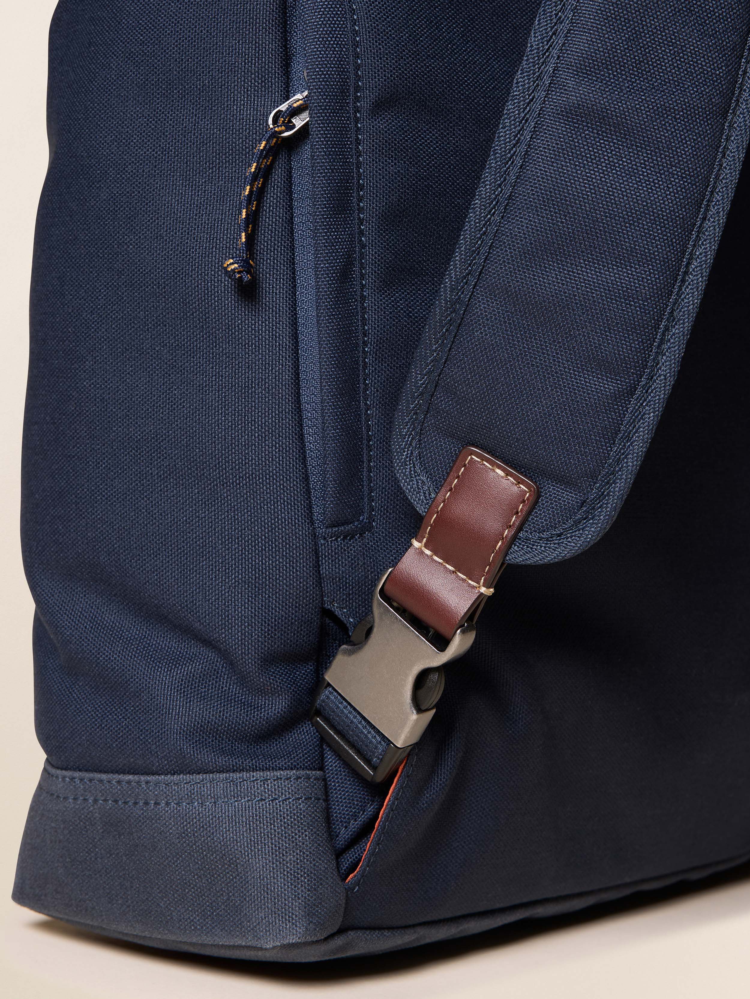 Close-up of a navy blue backpack with a brown leather strap and metal buckle on a beige background.