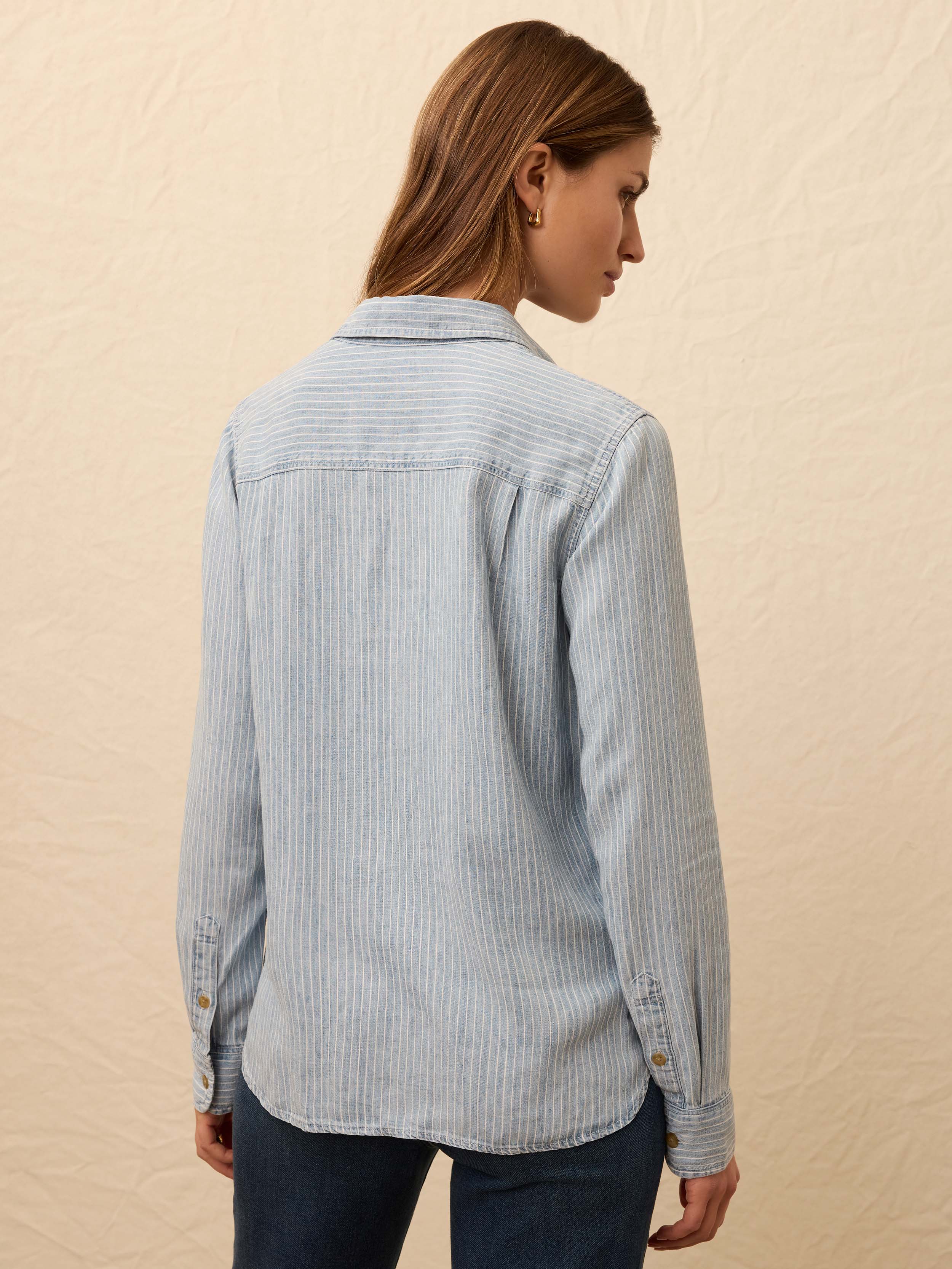 Woman wearing a light blue striped chambray shirt and dark blue jeans against a beige background