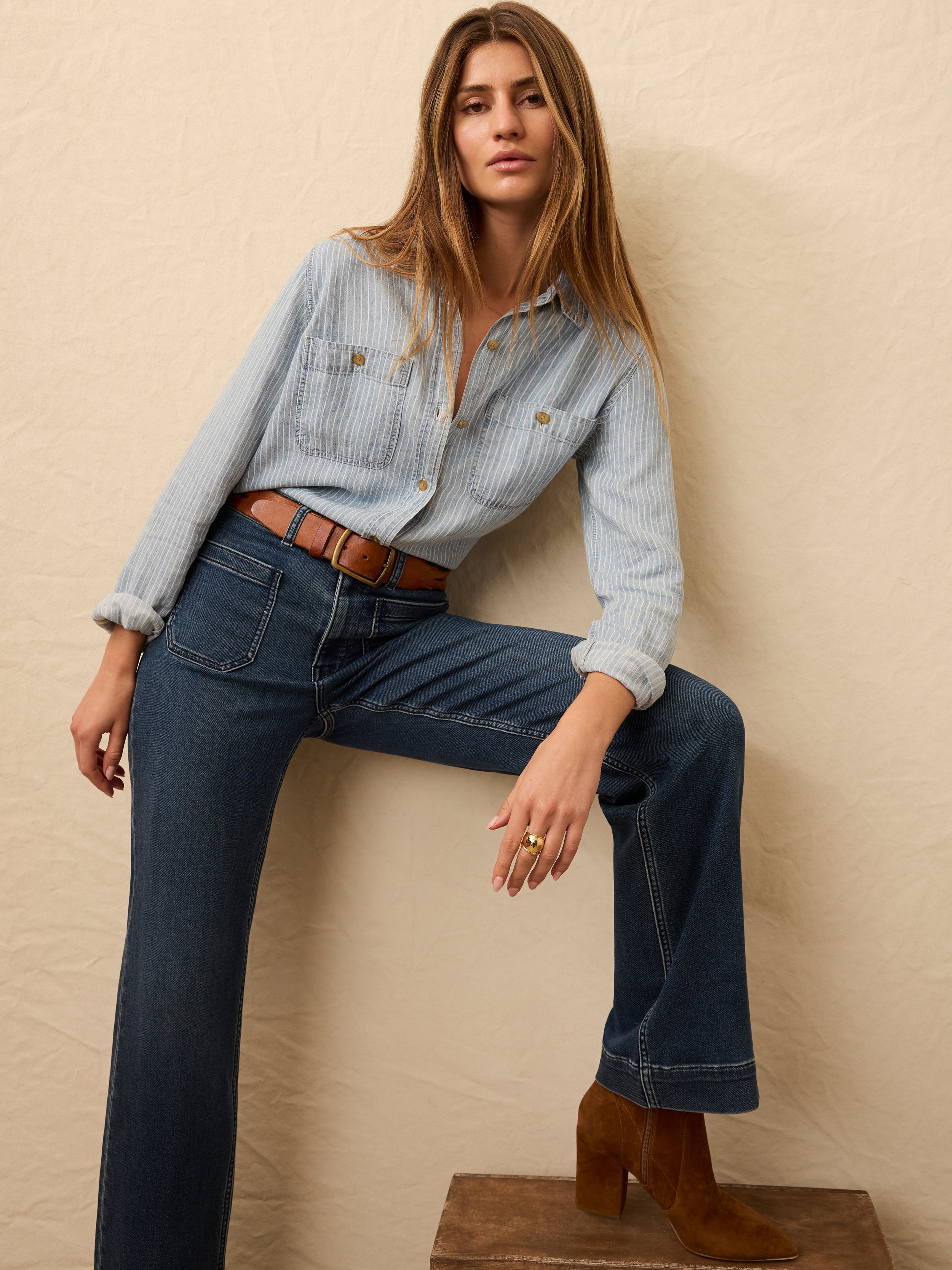Woman wearing a light blue striped chambray shirt and dark blue jeans against a beige background