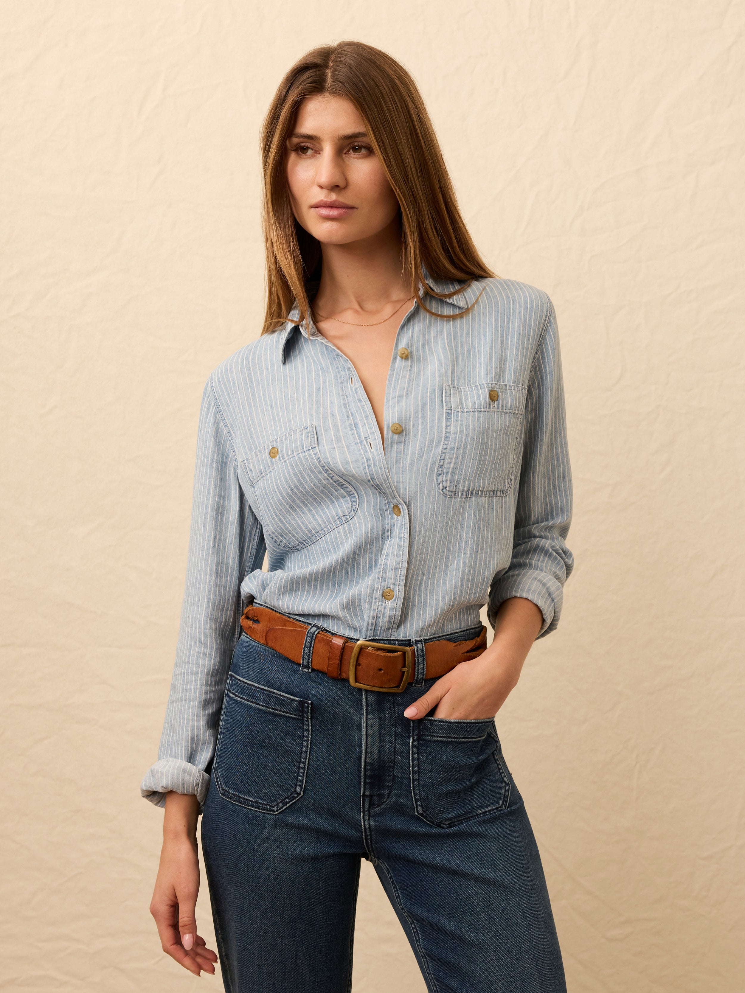 Woman wearing a light blue striped chambray shirt and dark blue jeans against a beige background