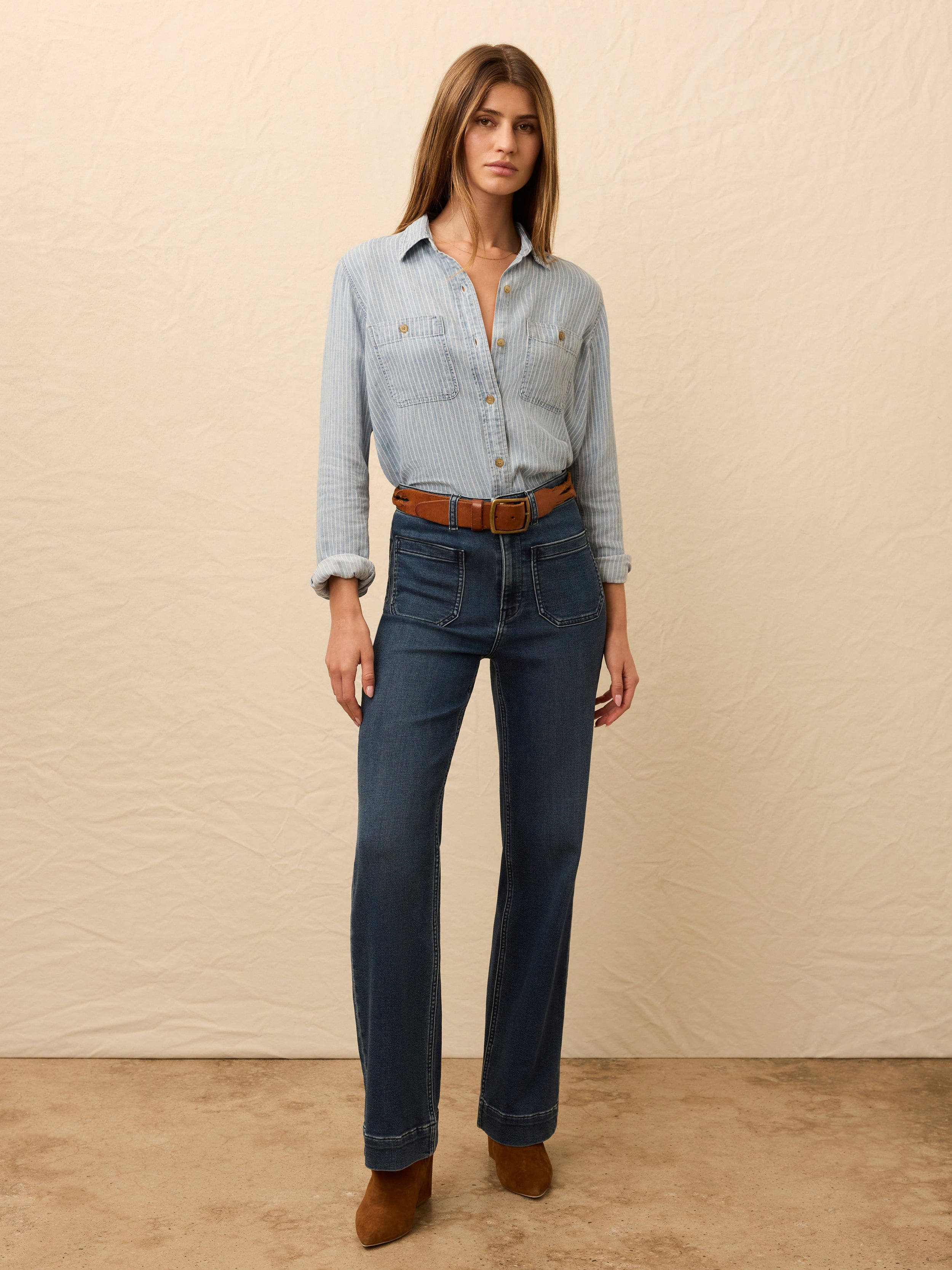 Woman wearing a light blue striped chambray shirt and dark blue jeans against a beige background