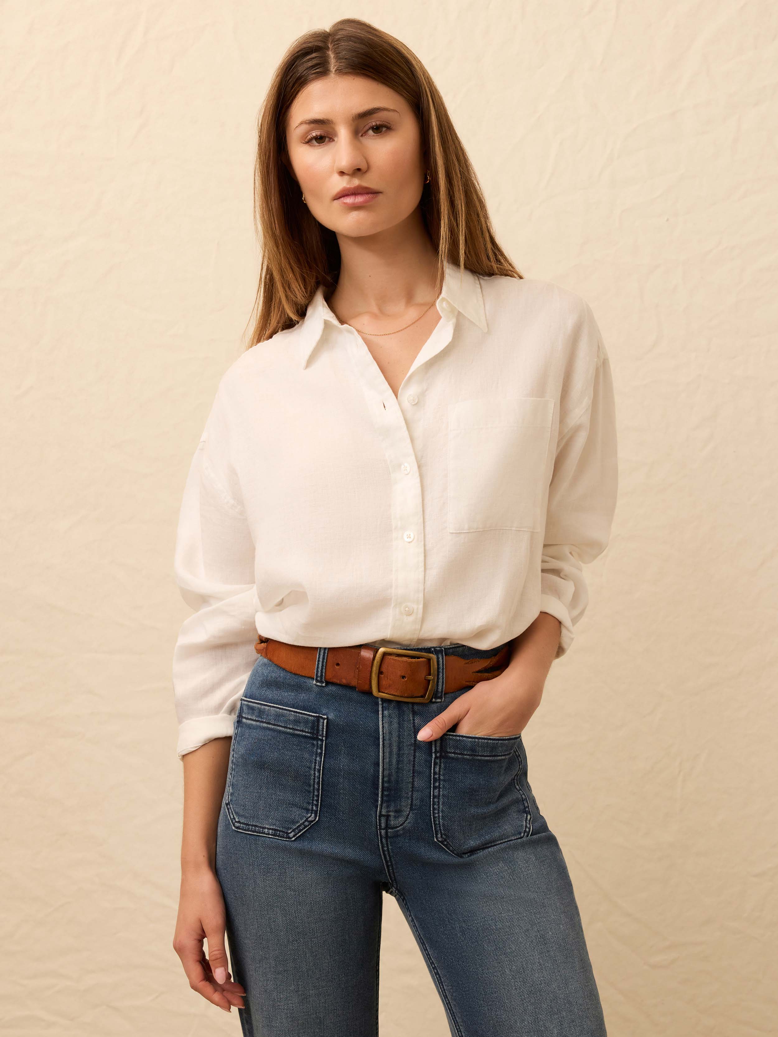 Woman wearing a white linen button up shirt and blue jeans with a brown belt against a beige background