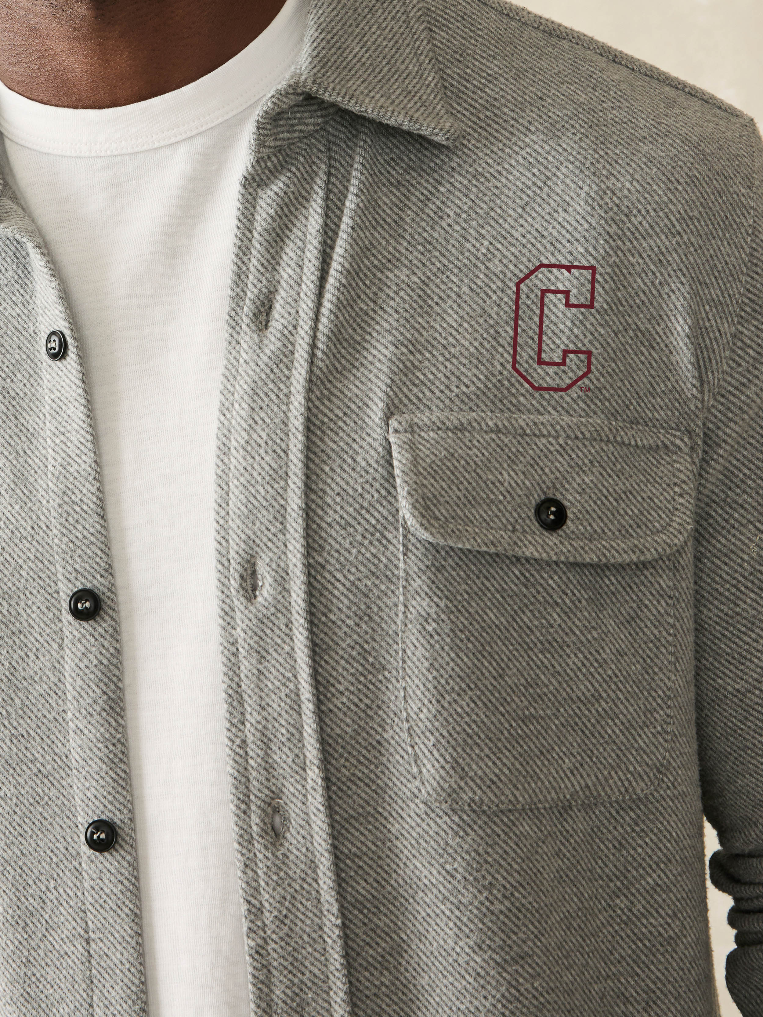 Man is wearing a grey long sleeve button down shirt with College of Charleston graphic on the left chest against a beige background