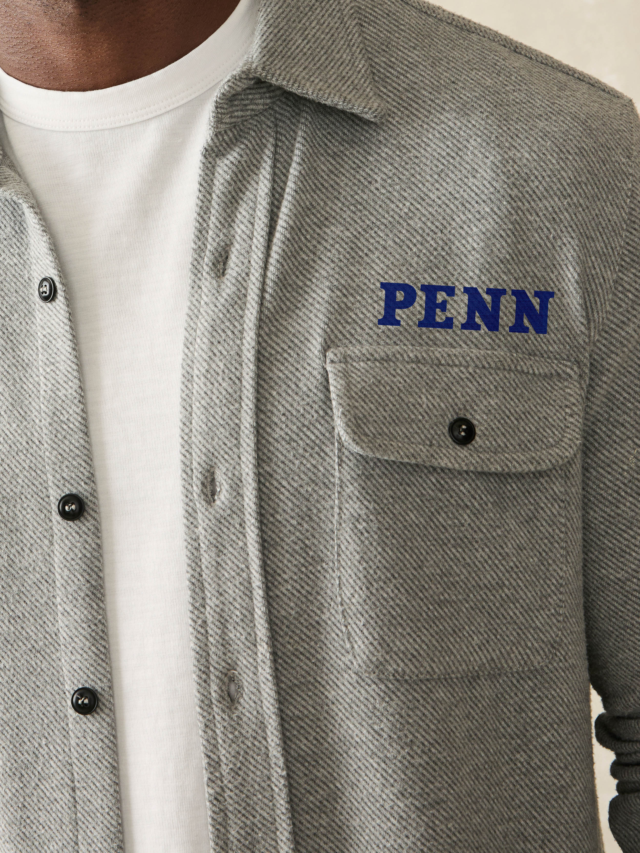 Man wearing a grey long sleeve button up shirt with University of Pennsylvania graphic on left chest and left arm against a beige background