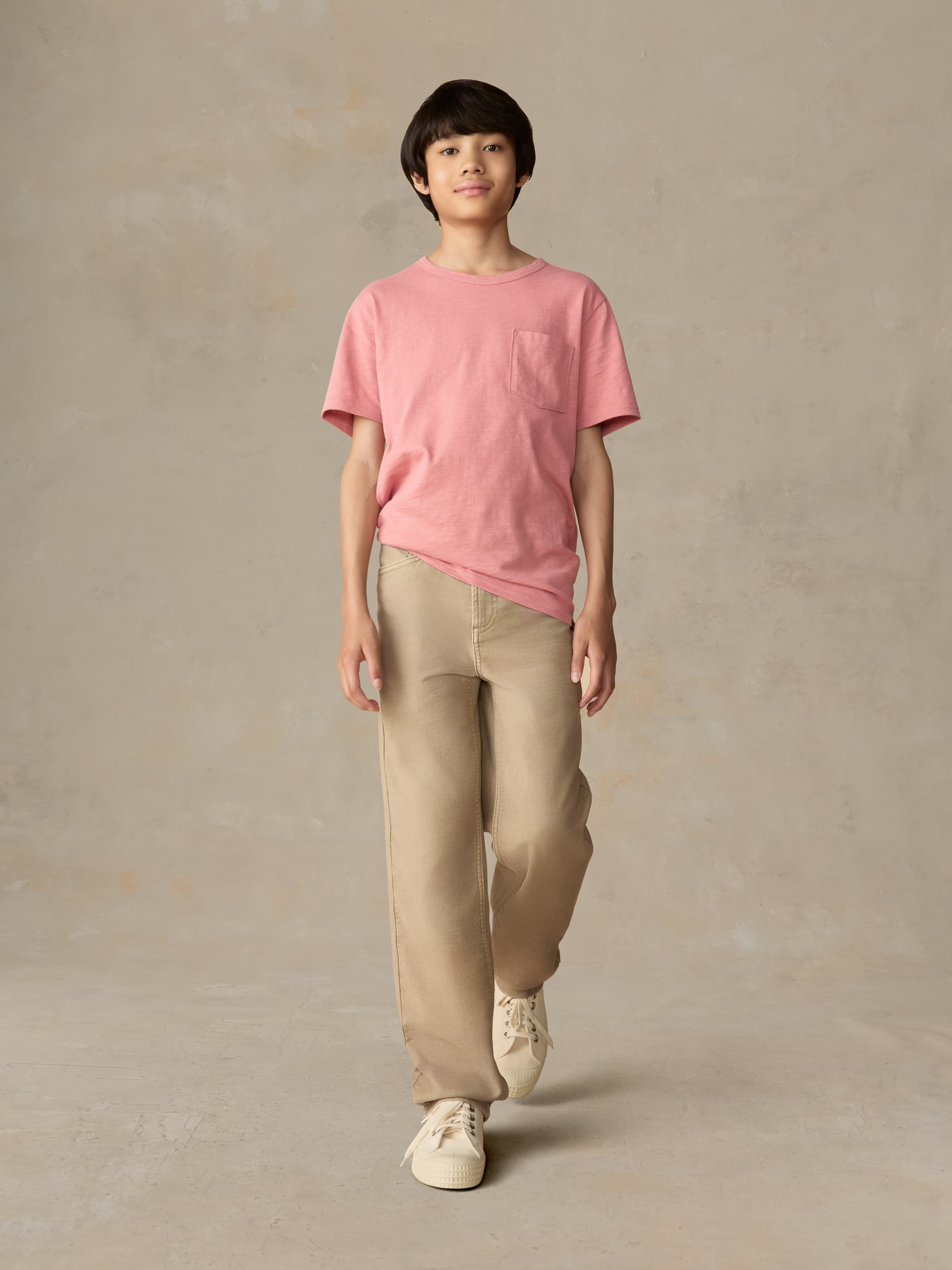 Child wearing a red pocket tee and beige pants against a beige background
