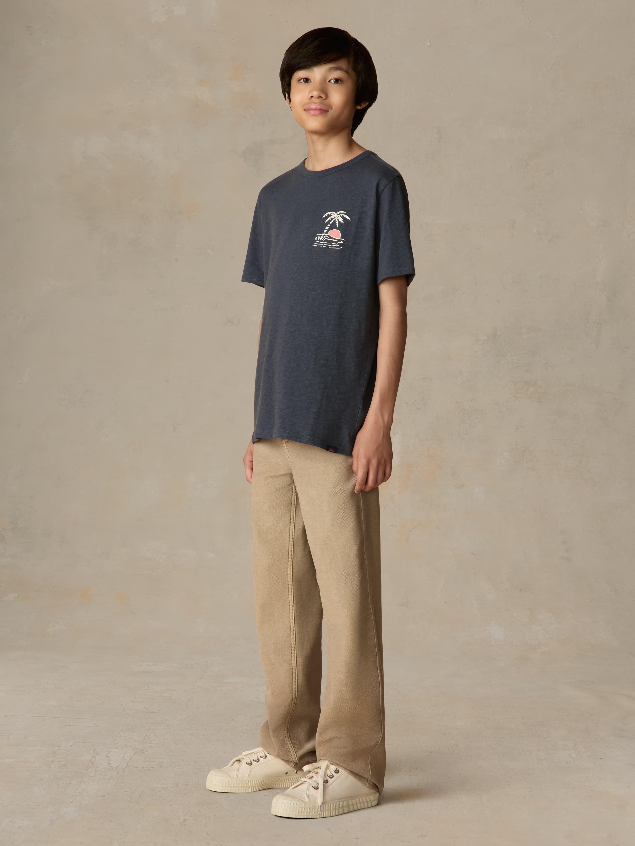 Child wearing a blue graphic shirt and beige pants against a beige background