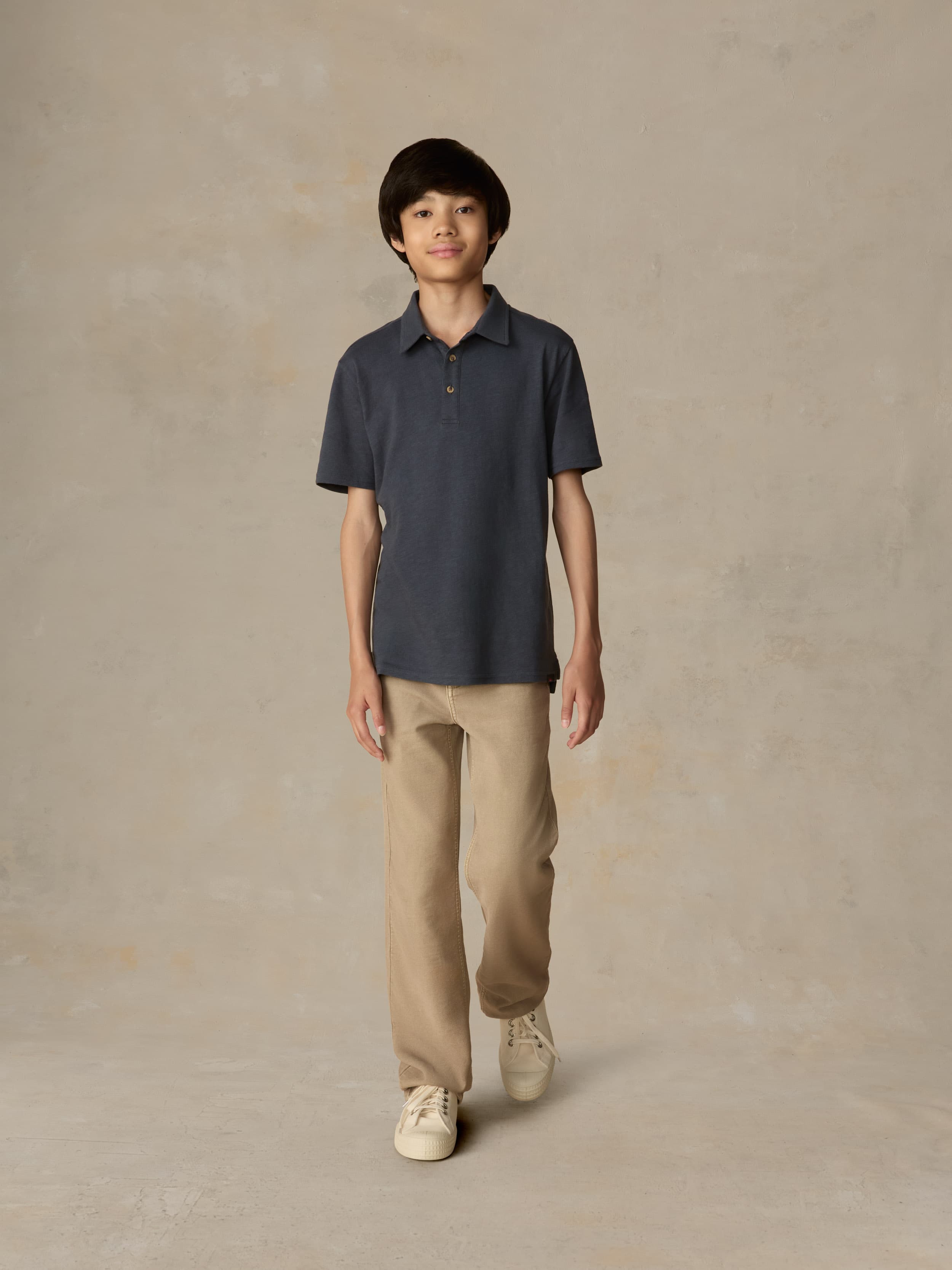 Child wearing a blue polo shirt and beige pants against a beige background