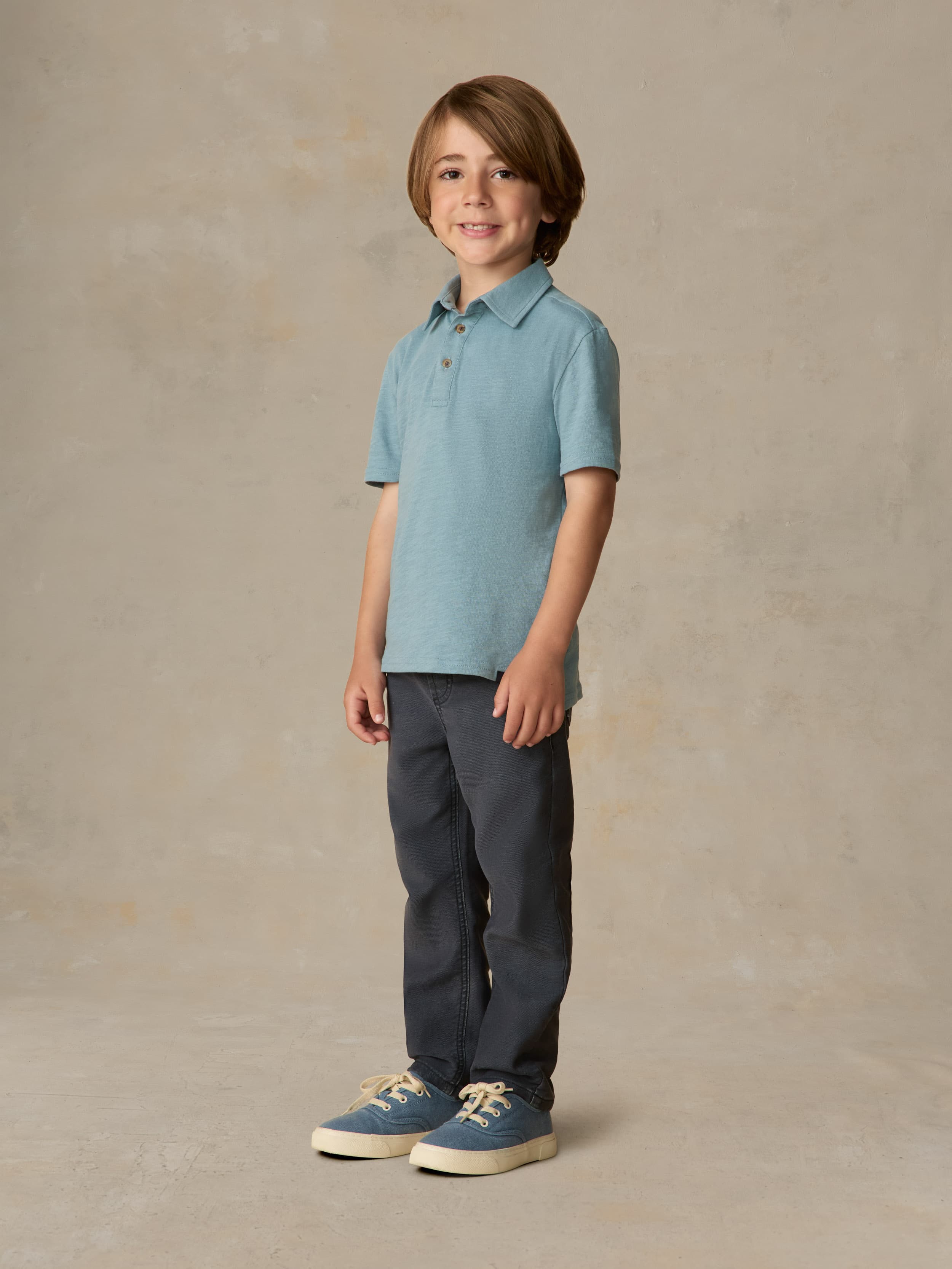 Child wearing a light blue polo shirt and navy pants against a beige background