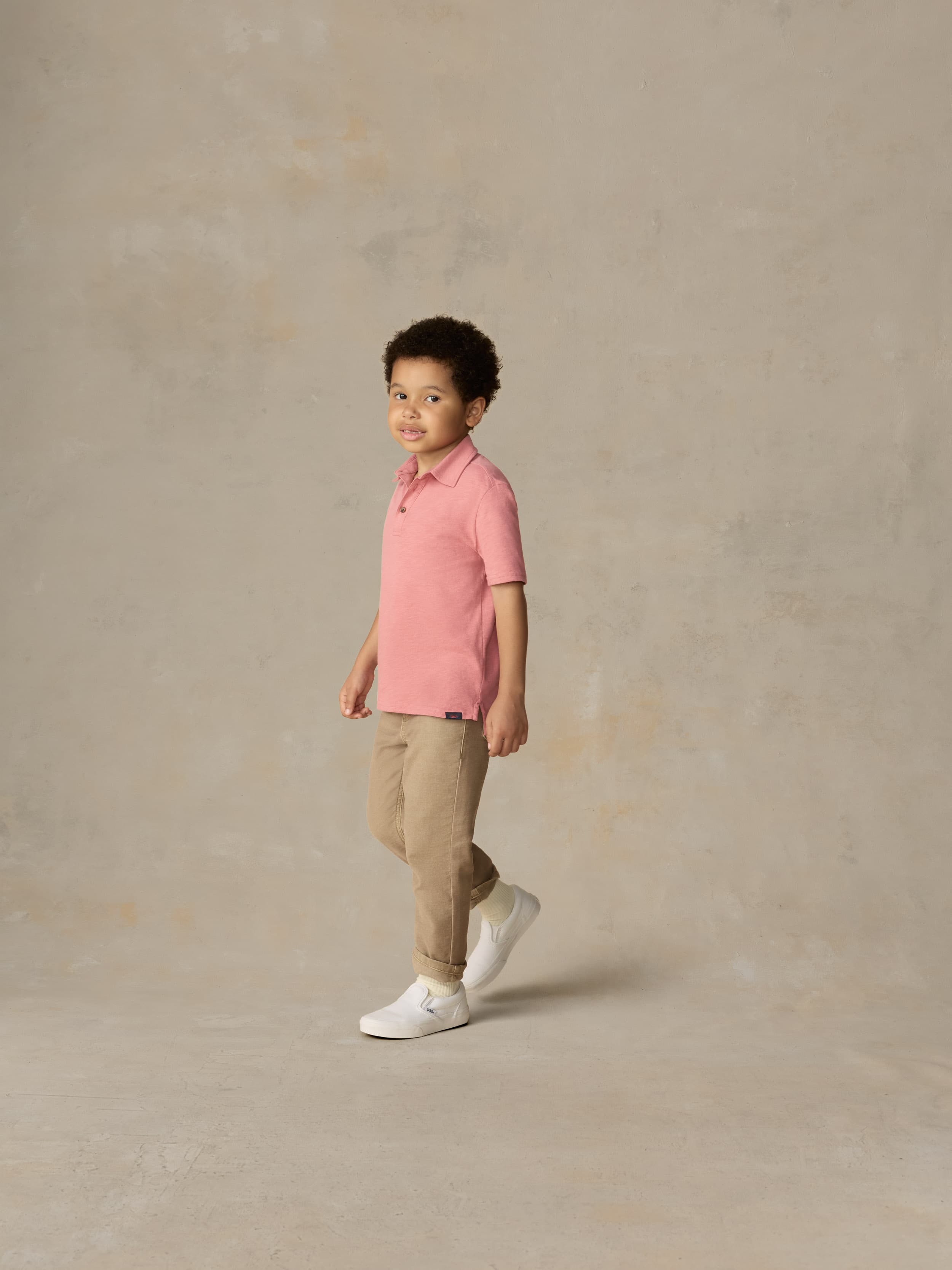 Child wearing a red polo shirt and beige pants against a beige background