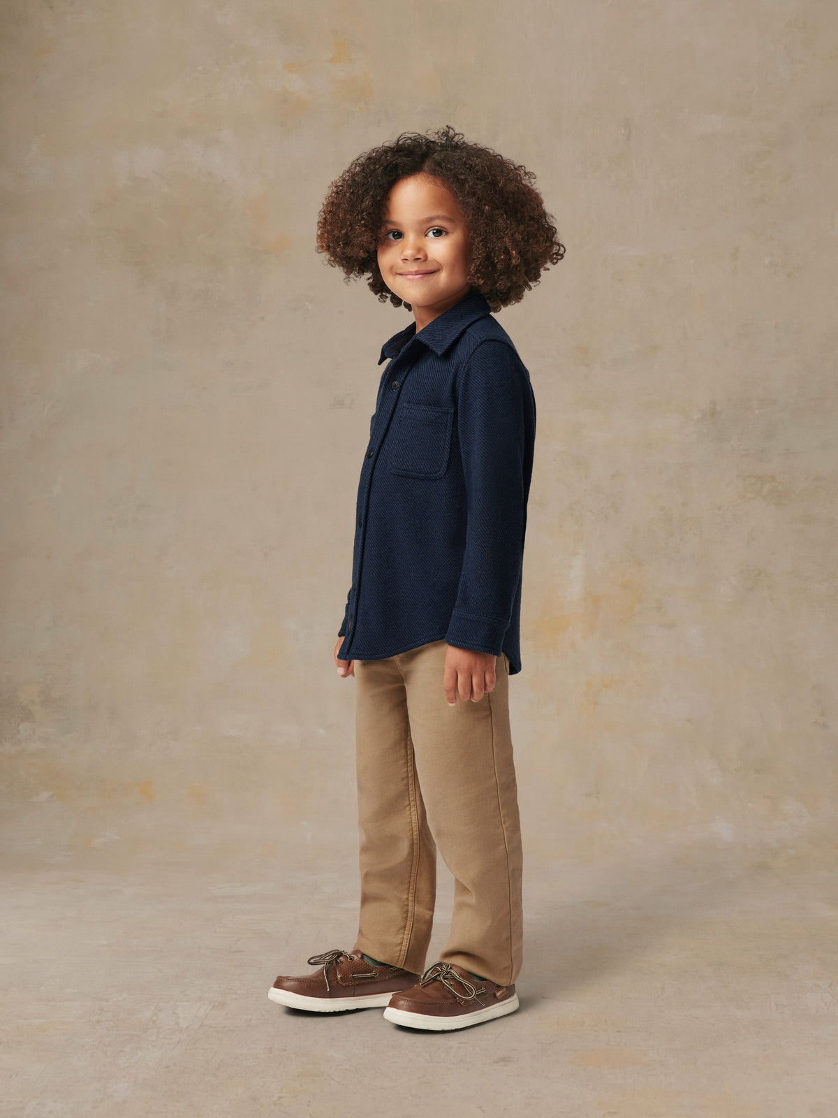 A child wearing a blue button up shirt and beige pants, image 5 of 6