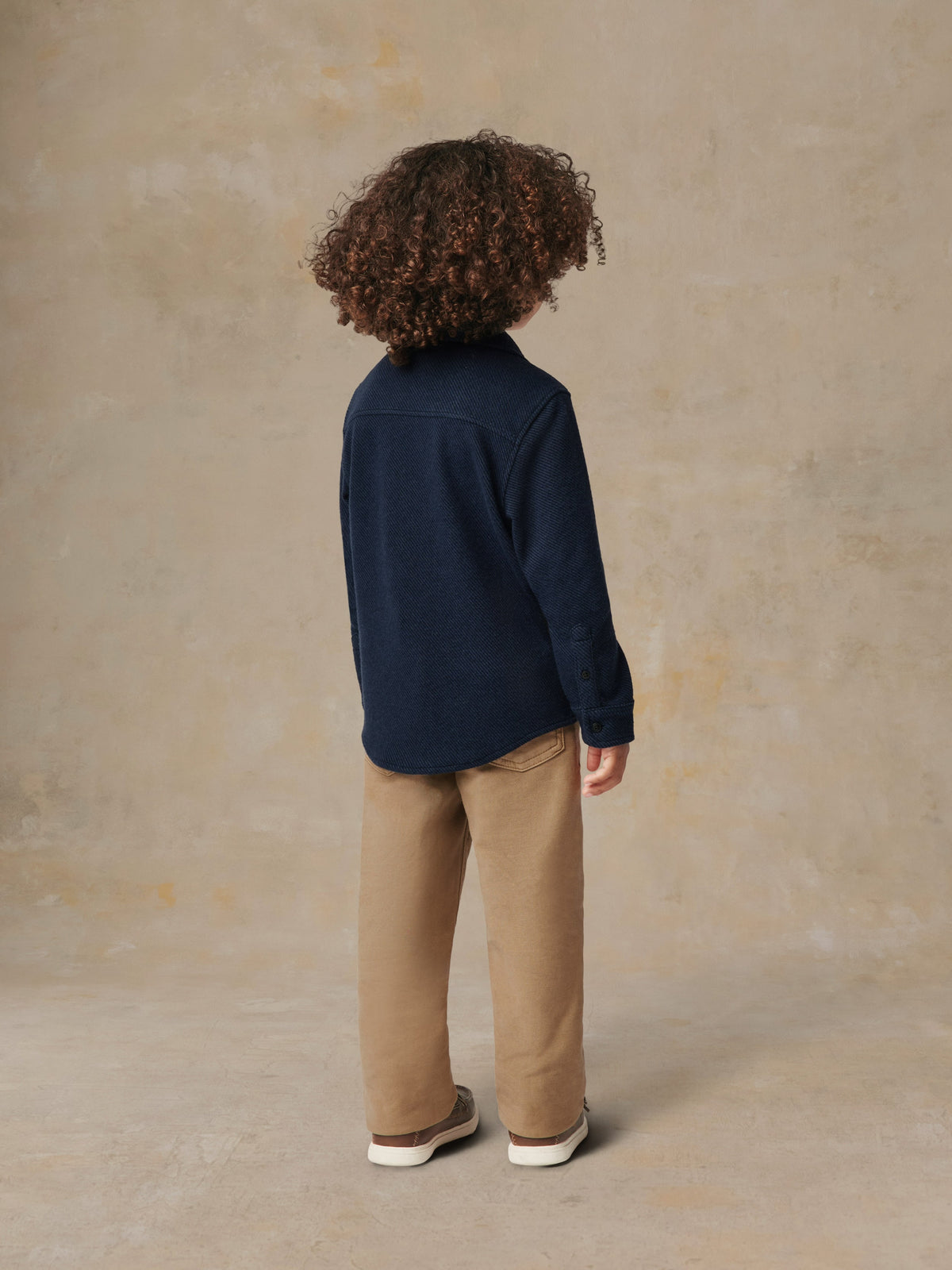 A child viewed from behind wearing a blue button up shirt and beige pants, image 3 of 6