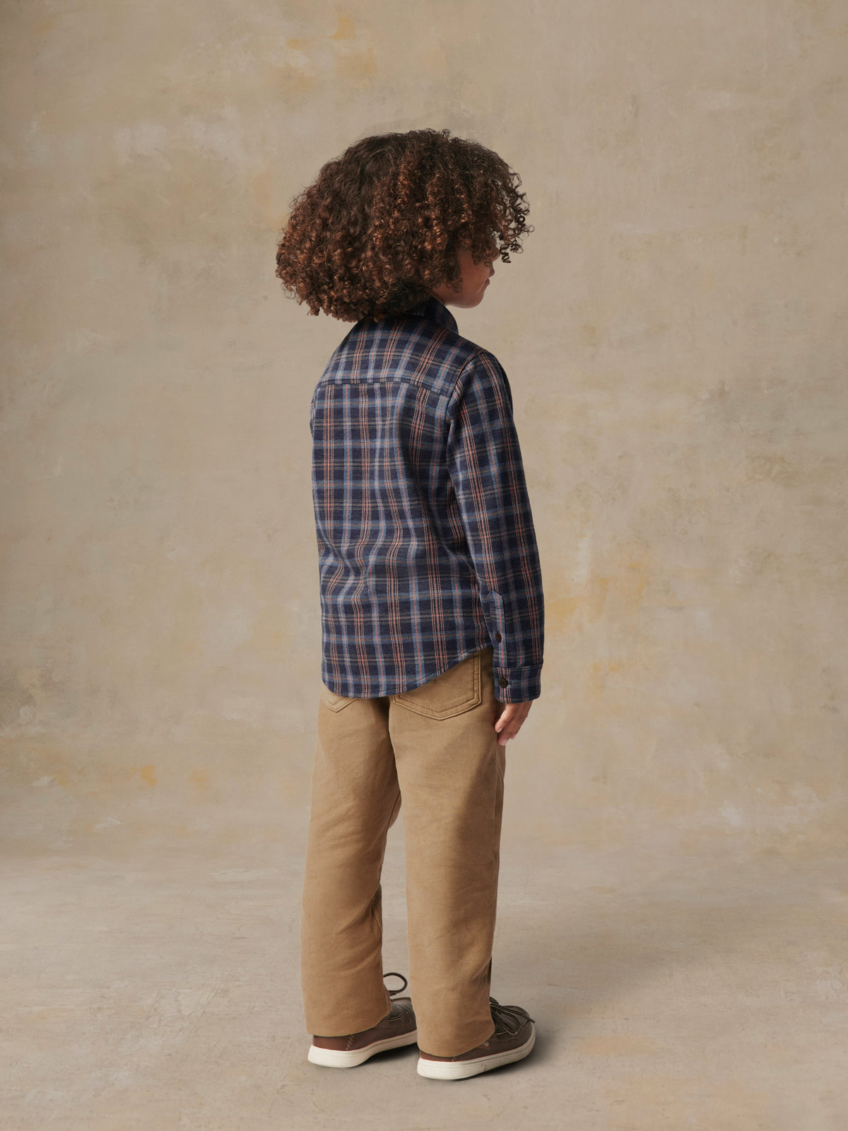 A child viewed from behind wearing a plaid shirt and beige pants, image 4 of 6