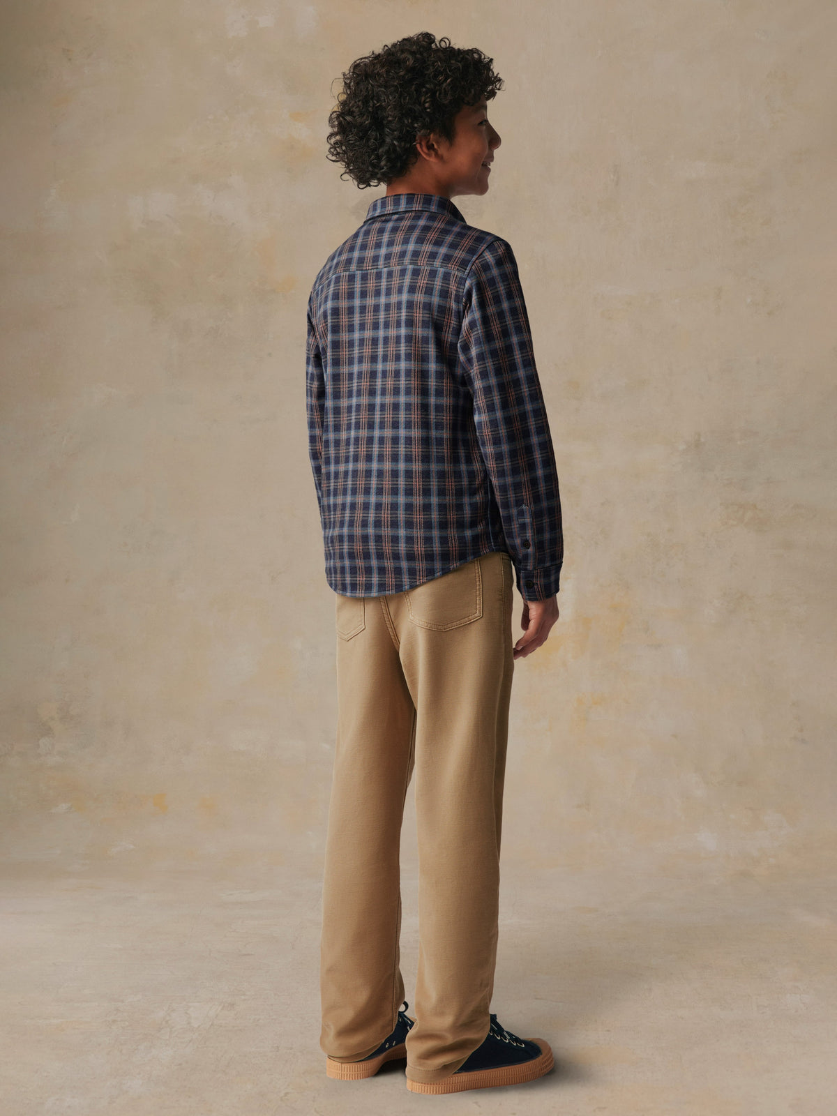A child viewed from behind wearing a plaid button down shirt and beige pants, image 5 of 7